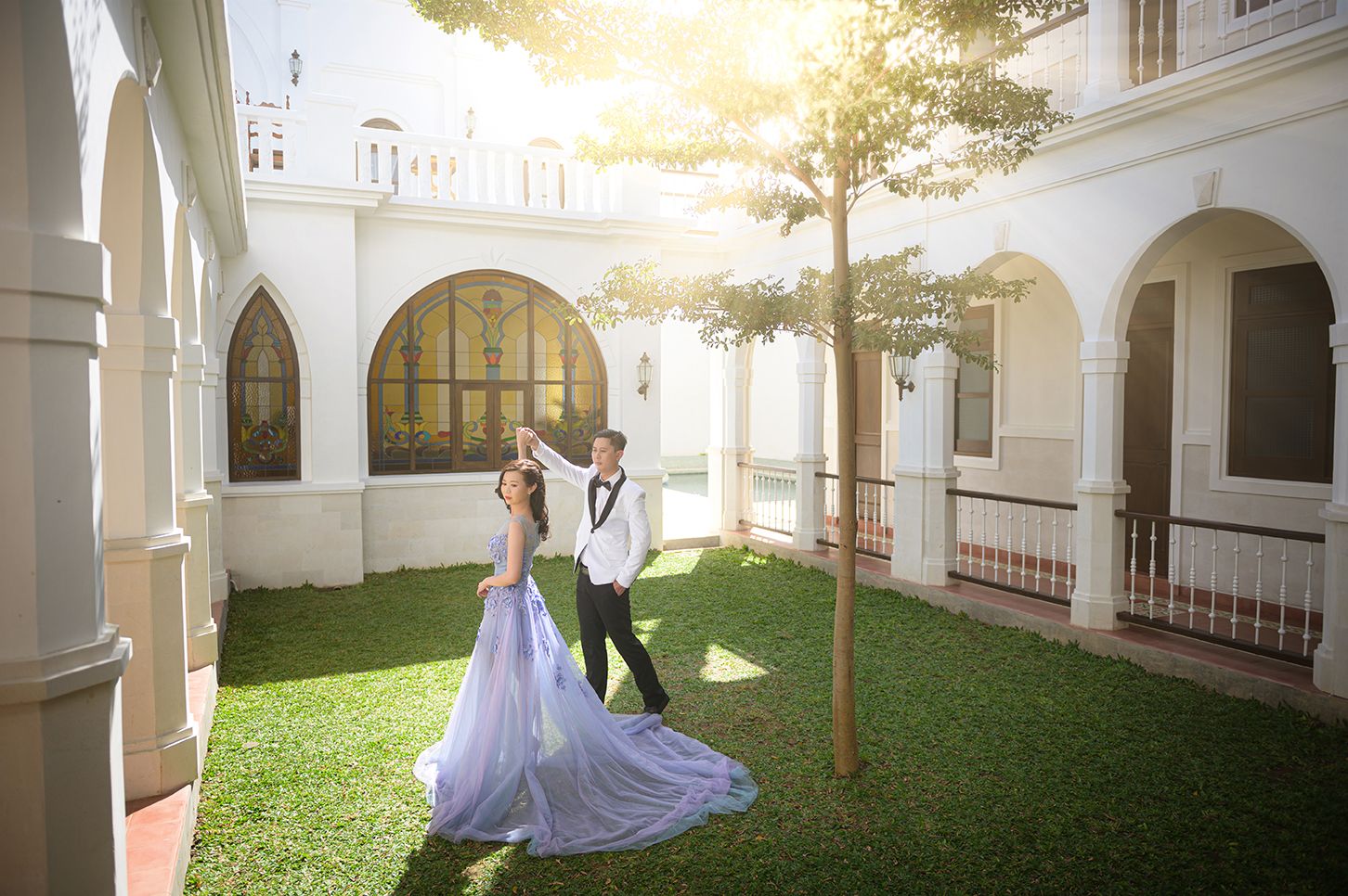 Couple in wedding attire posing on green lawn outside elegant white colonial building with arched windows and sunlight.
