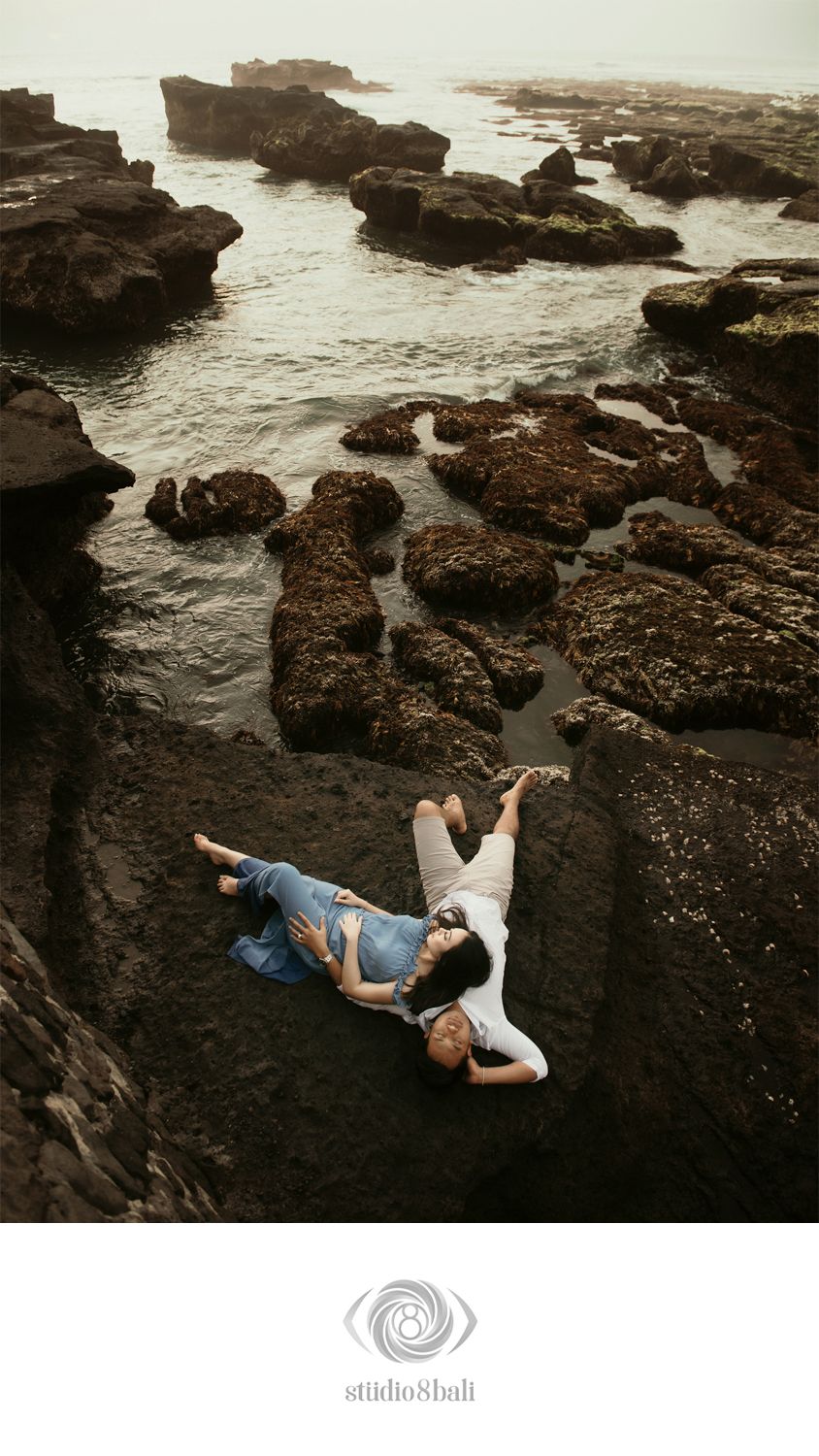 Couple lying on rocky coastal shore in Bali, surrounded by tide pools and ocean waves, captured by Studio8Bali.