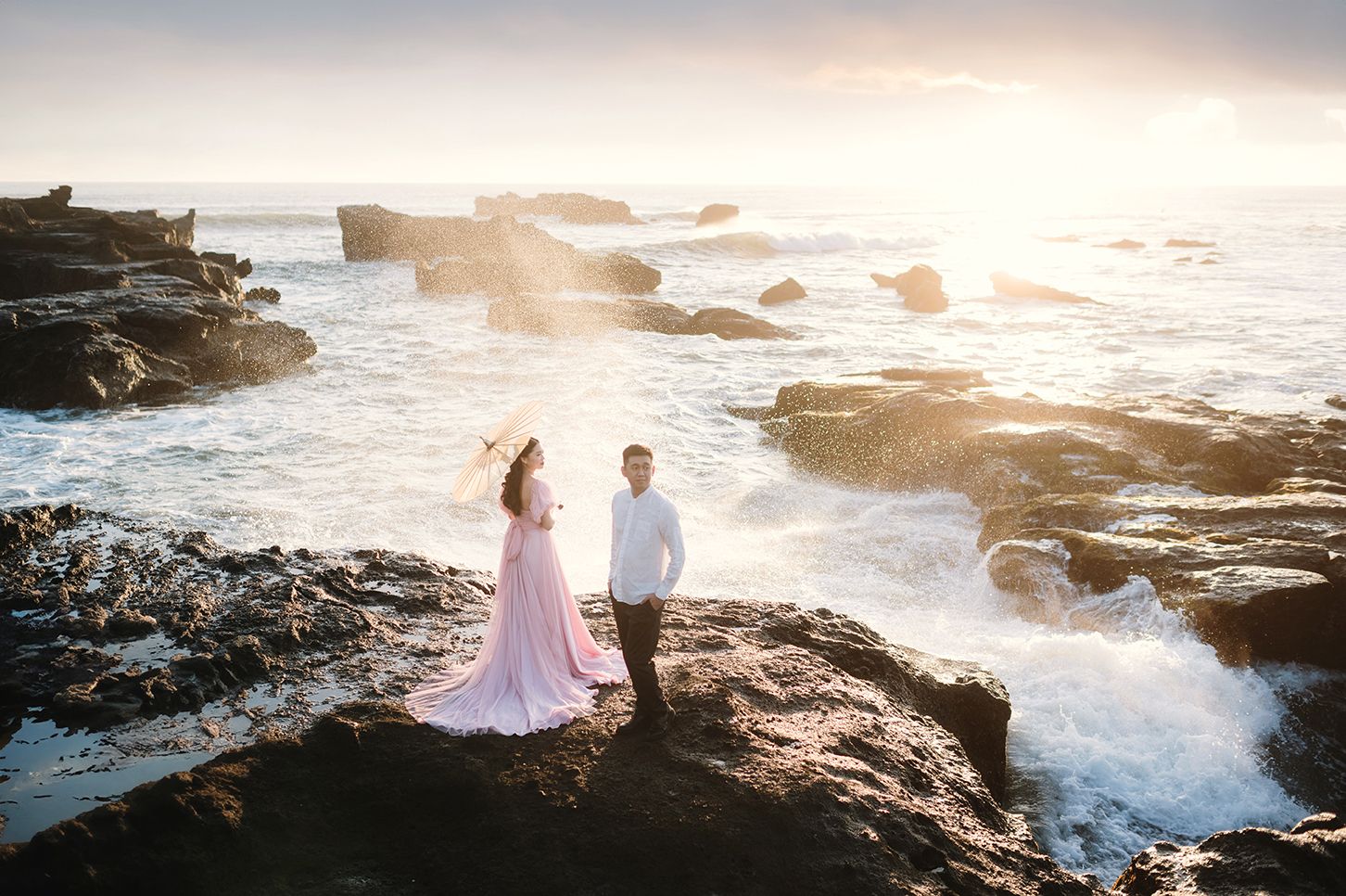 Couple poses on rocky ocean shore at sunset, woman in pink flowing dress, waves crashing behind them.