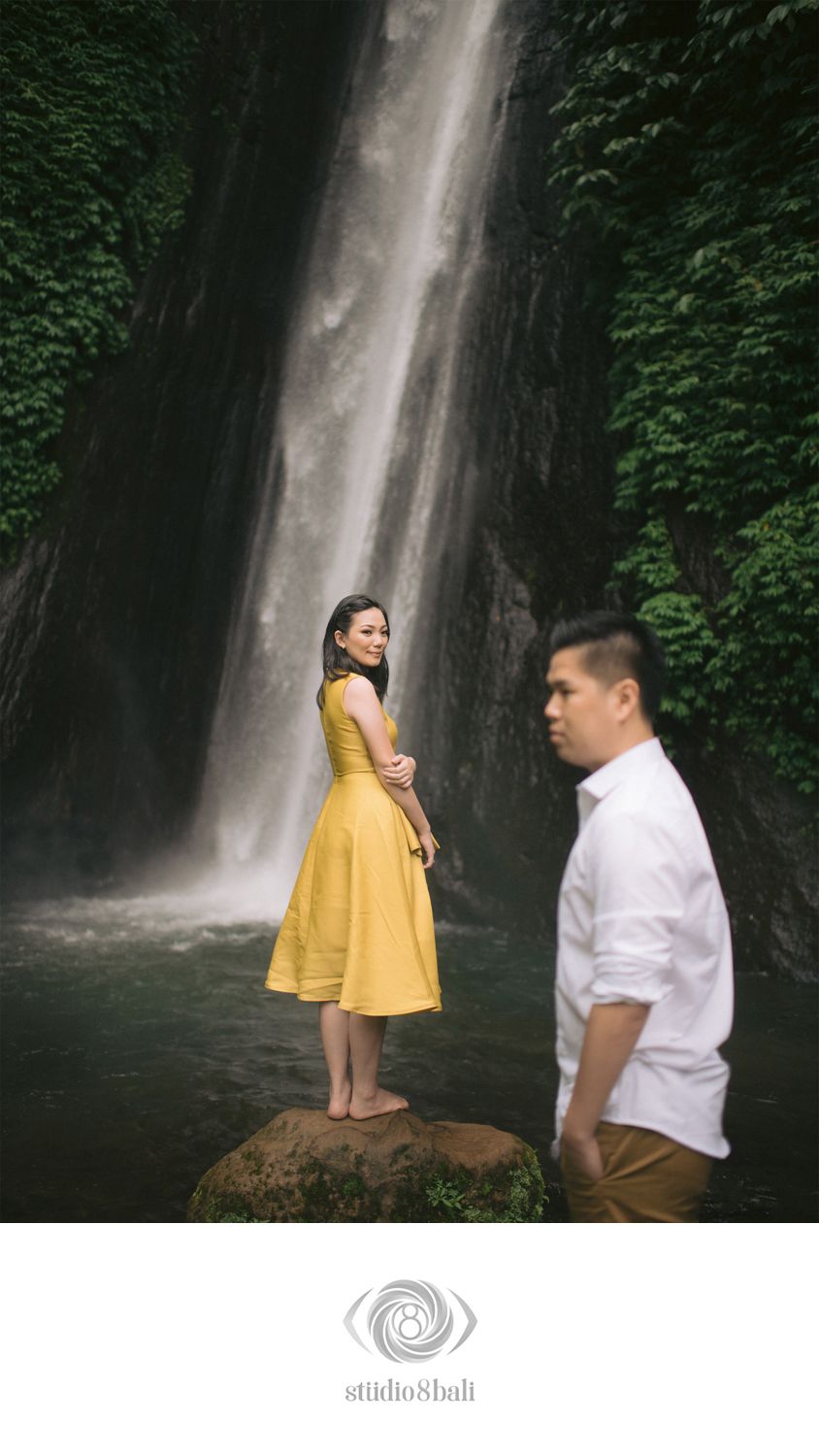Couple poses near a lush waterfall in Bali, woman in yellow dress stands on rock as man looks away.