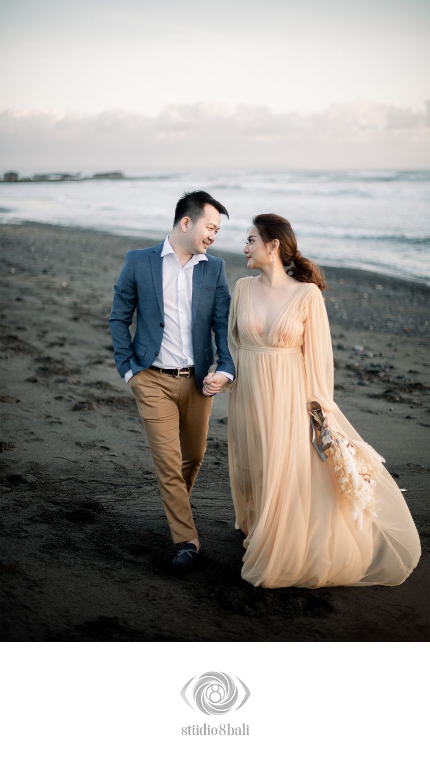 Couple walking hand in hand on a beach, man in blue blazer, woman in flowing peach gown, smiling at each other.