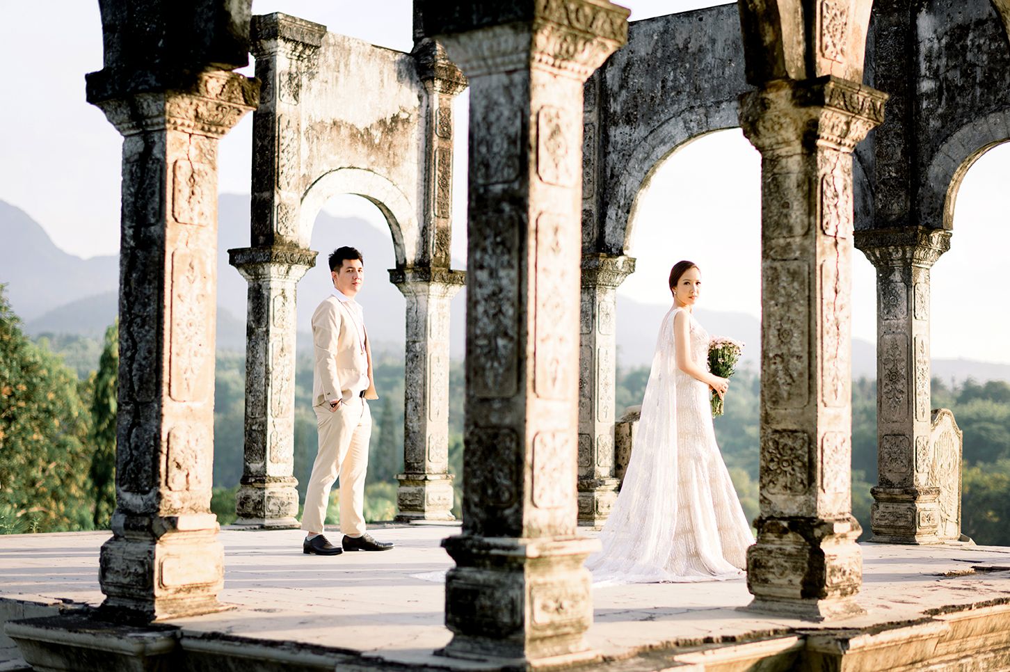 Bride and groom pose among ancient stone arches and columns with mountain views in a romantic outdoor wedding photo.