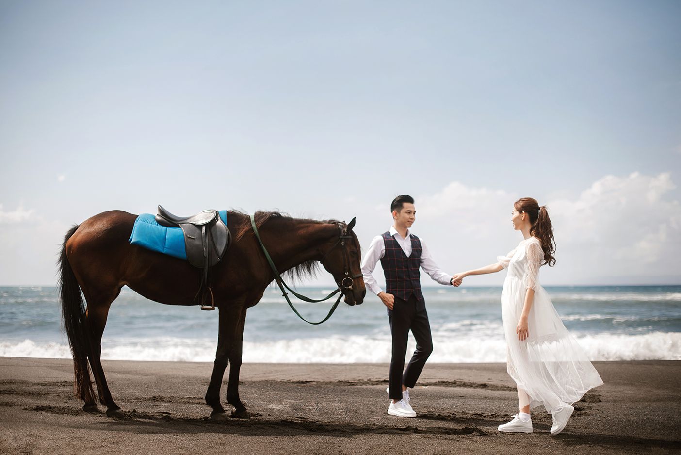Couple in wedding attire holding hands beside a brown horse on a scenic black sand beach.