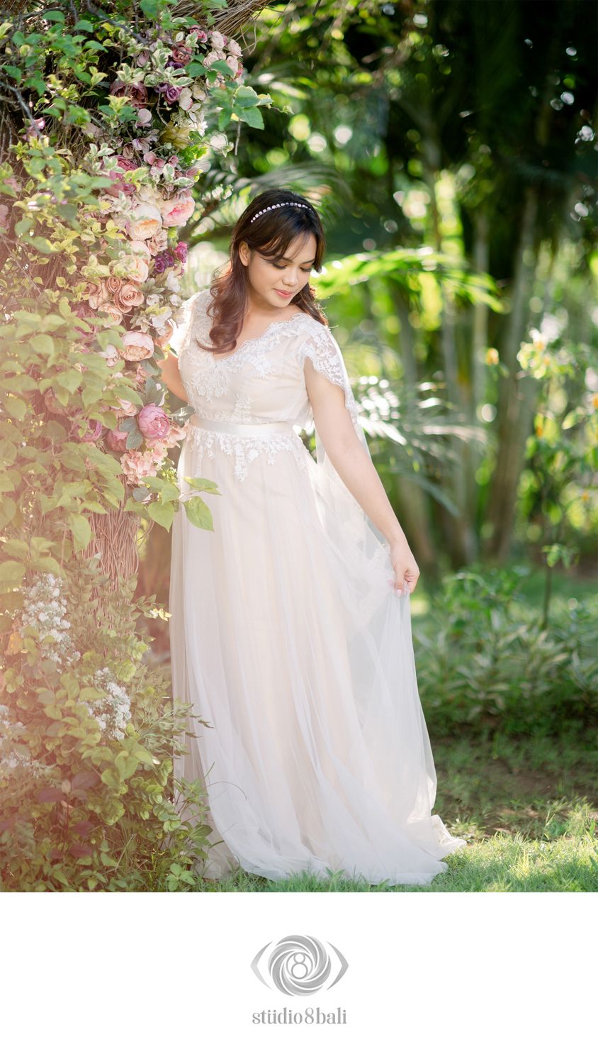 Bride in lace wedding gown poses among blooming flowers in a lush garden, wearing a delicate headband.