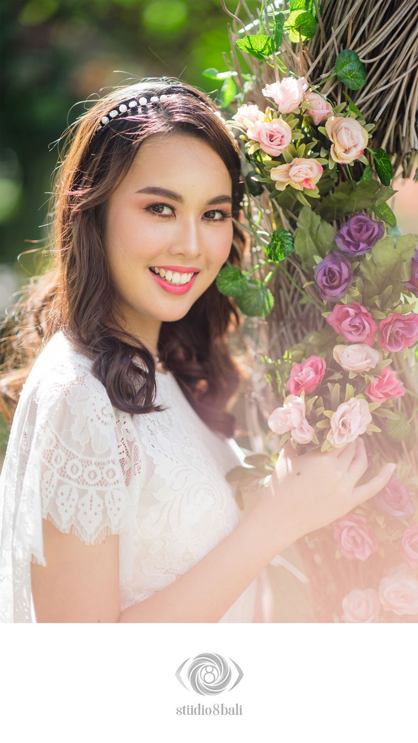 Smiling bride in white lace dress with pearl headband beside pink roses, Studio 8 Bali.