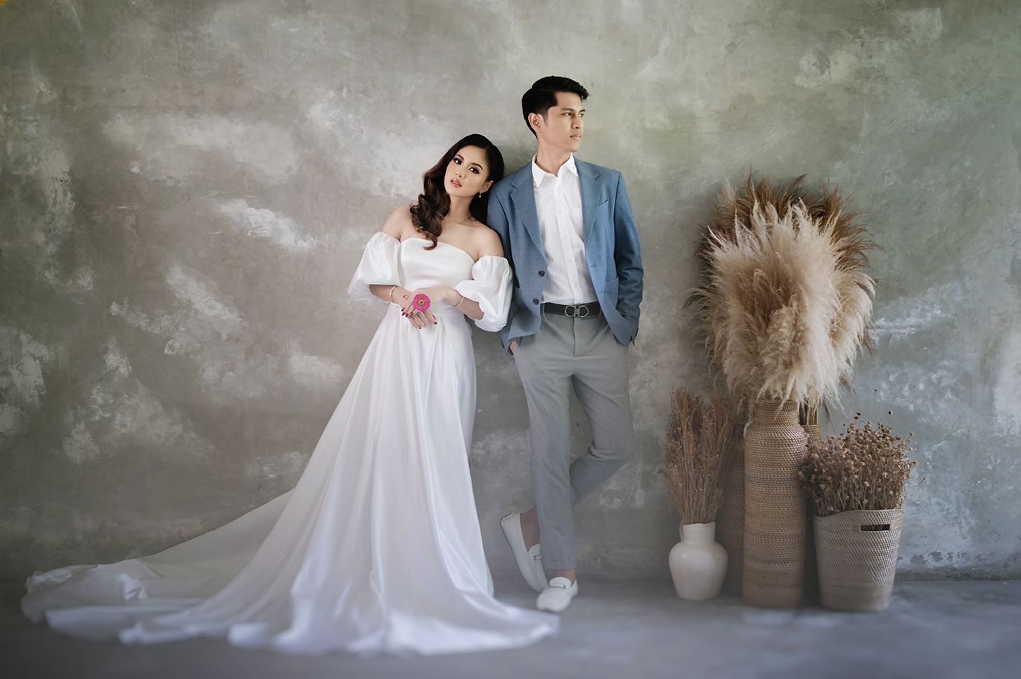 Elegant couple in wedding attire posing before a textured gray backdrop with pampas grass decor.