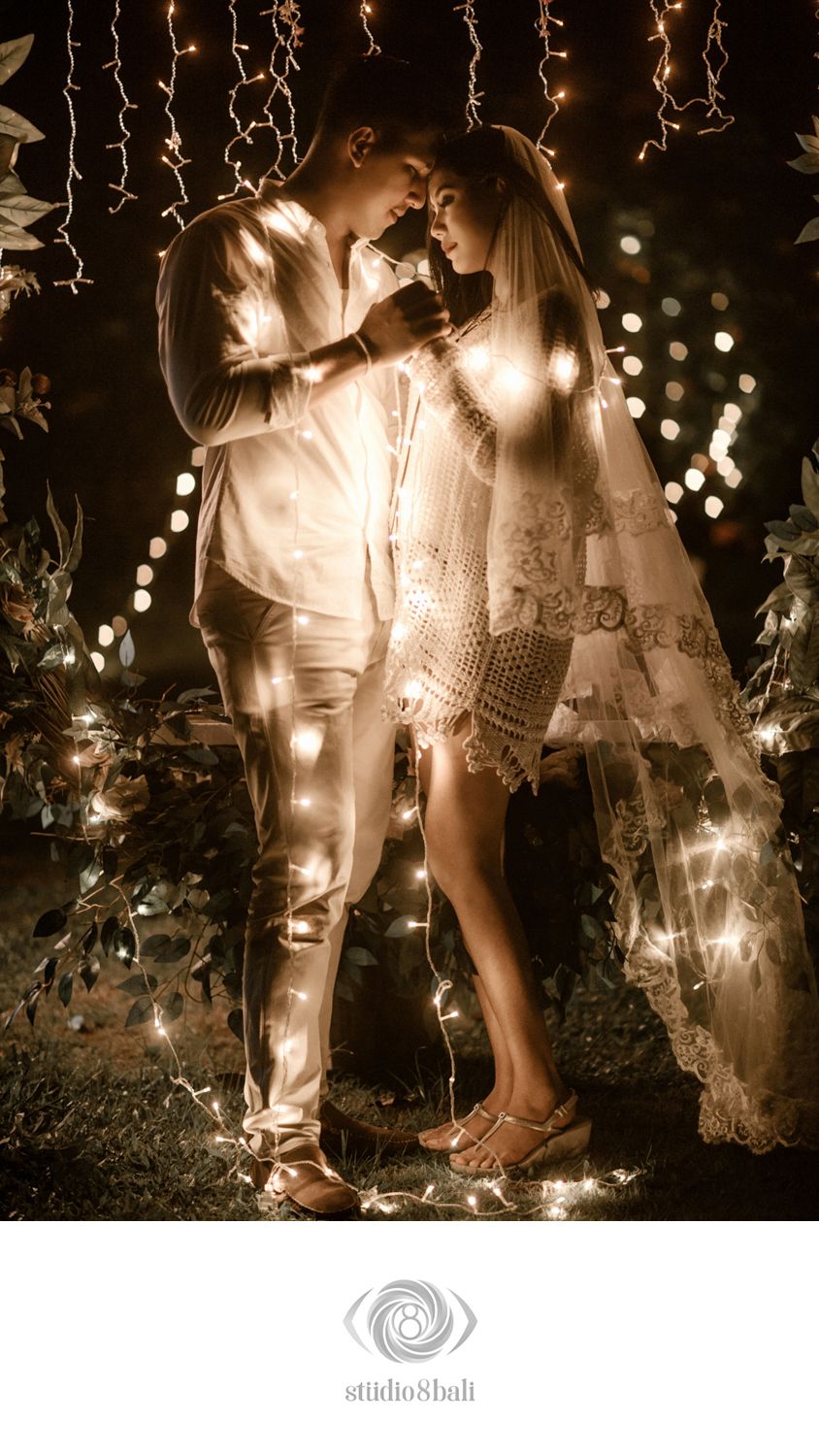 Romantic couple embracing under fairy lights at a Bali wedding, bride in lace dress with veil glowing in warm bokeh light.