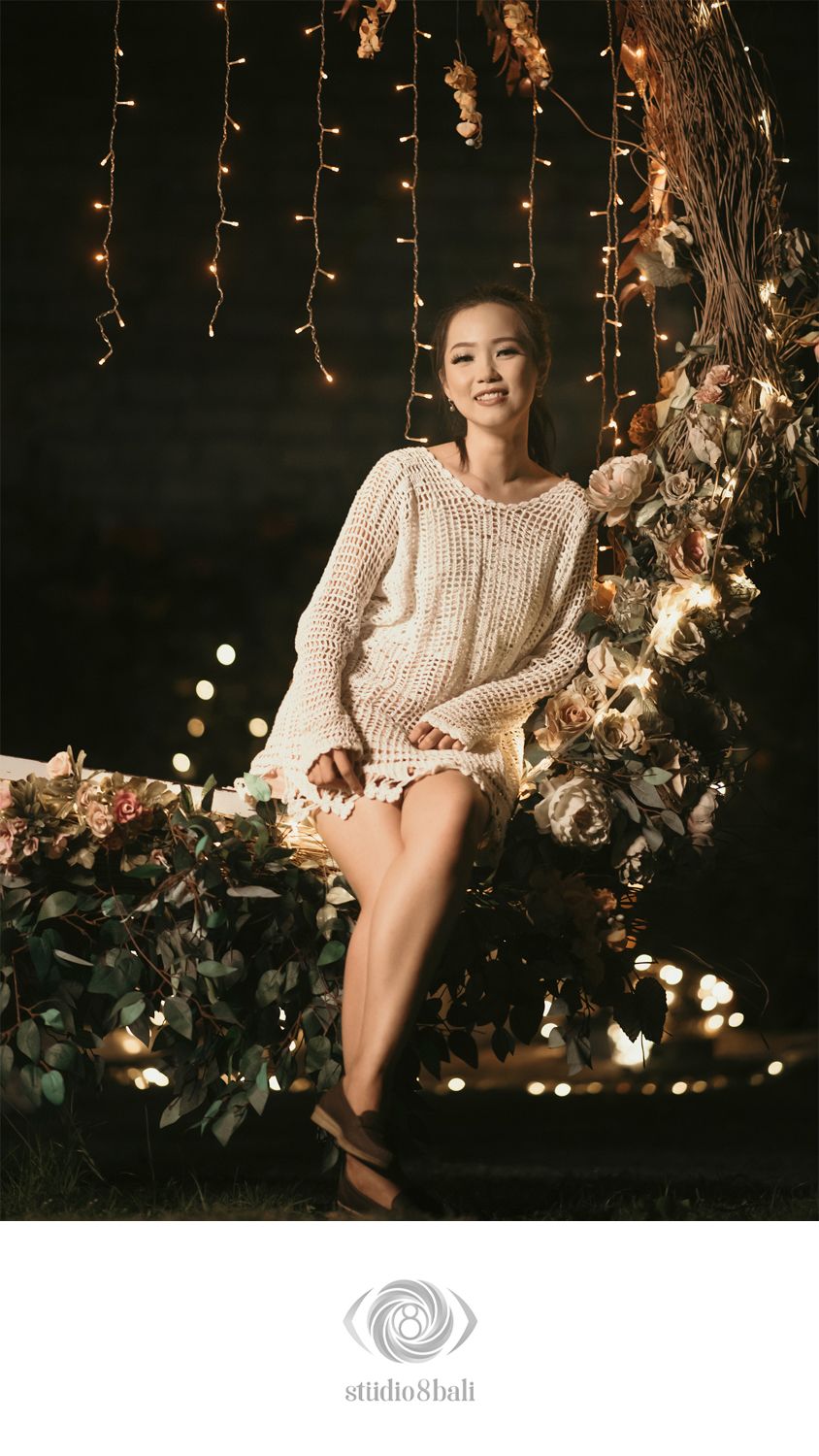 A smiling woman in a cream knit sweater sits on a floral swing surrounded by warm fairy lights at night.