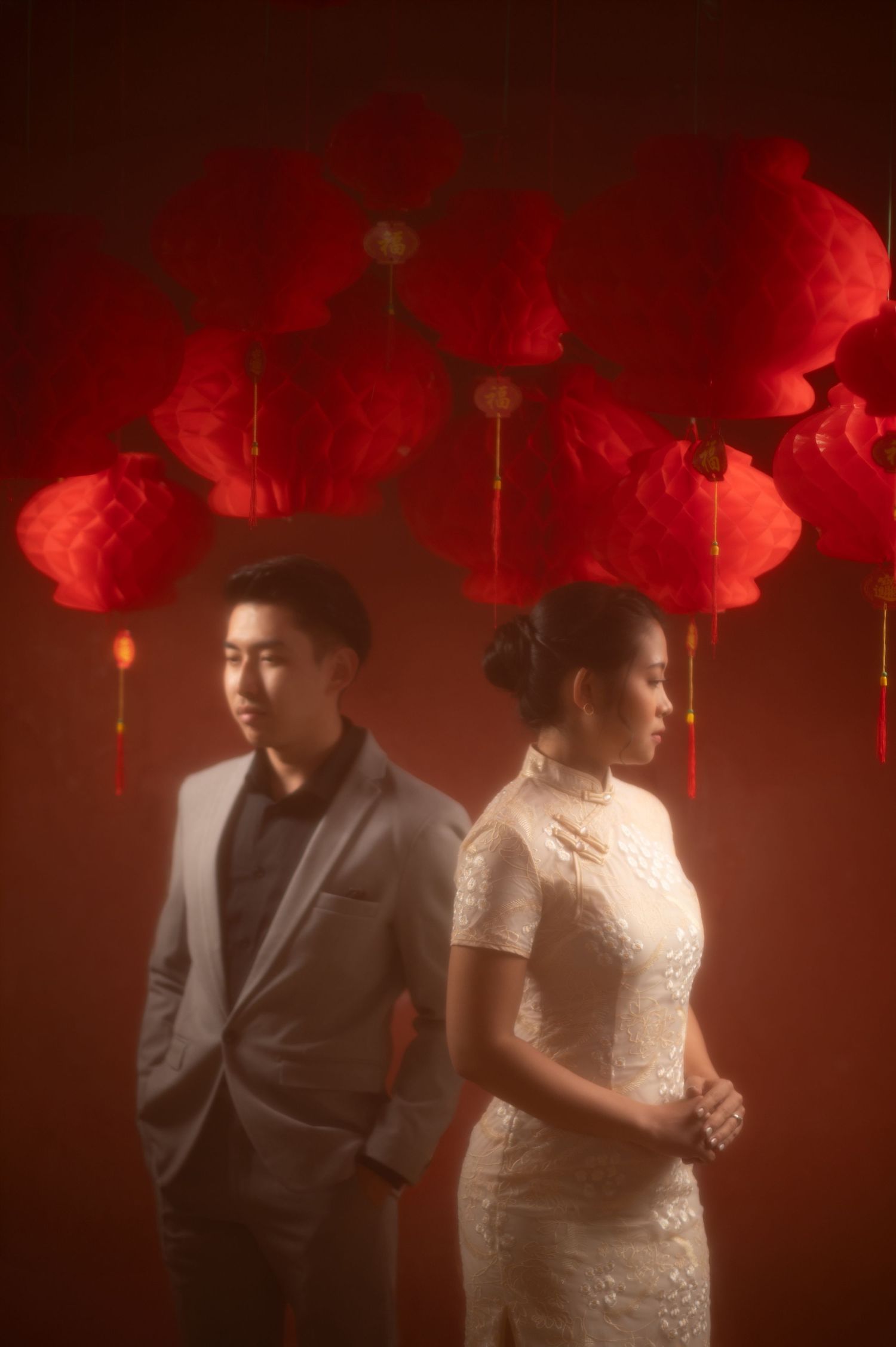 Couple in wedding attire posing under red Chinese lanterns, man in gray suit, woman in white lace qipao dress.