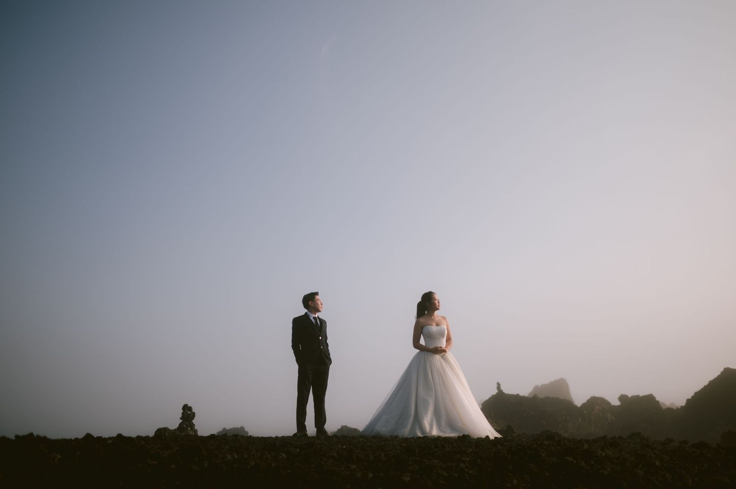 Bride in white gown and groom in black suit stand apart on rocky terrain at dusk with misty sky backdrop.