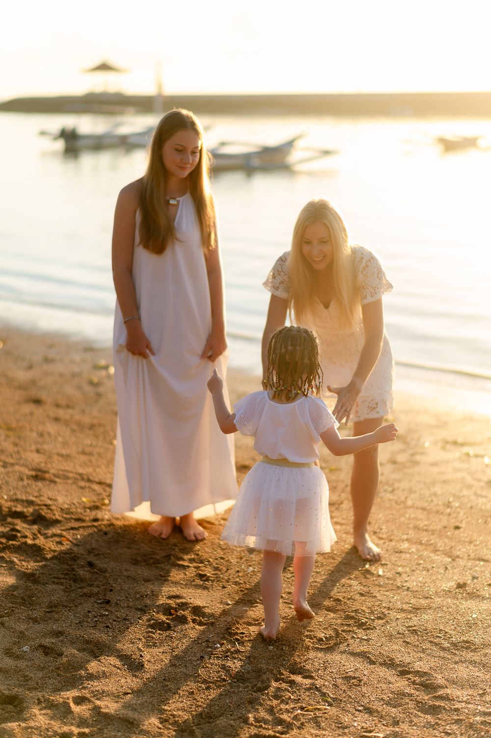 Blonde woman and teenage girl playing with young child in white dress on sandy beach at sunset.