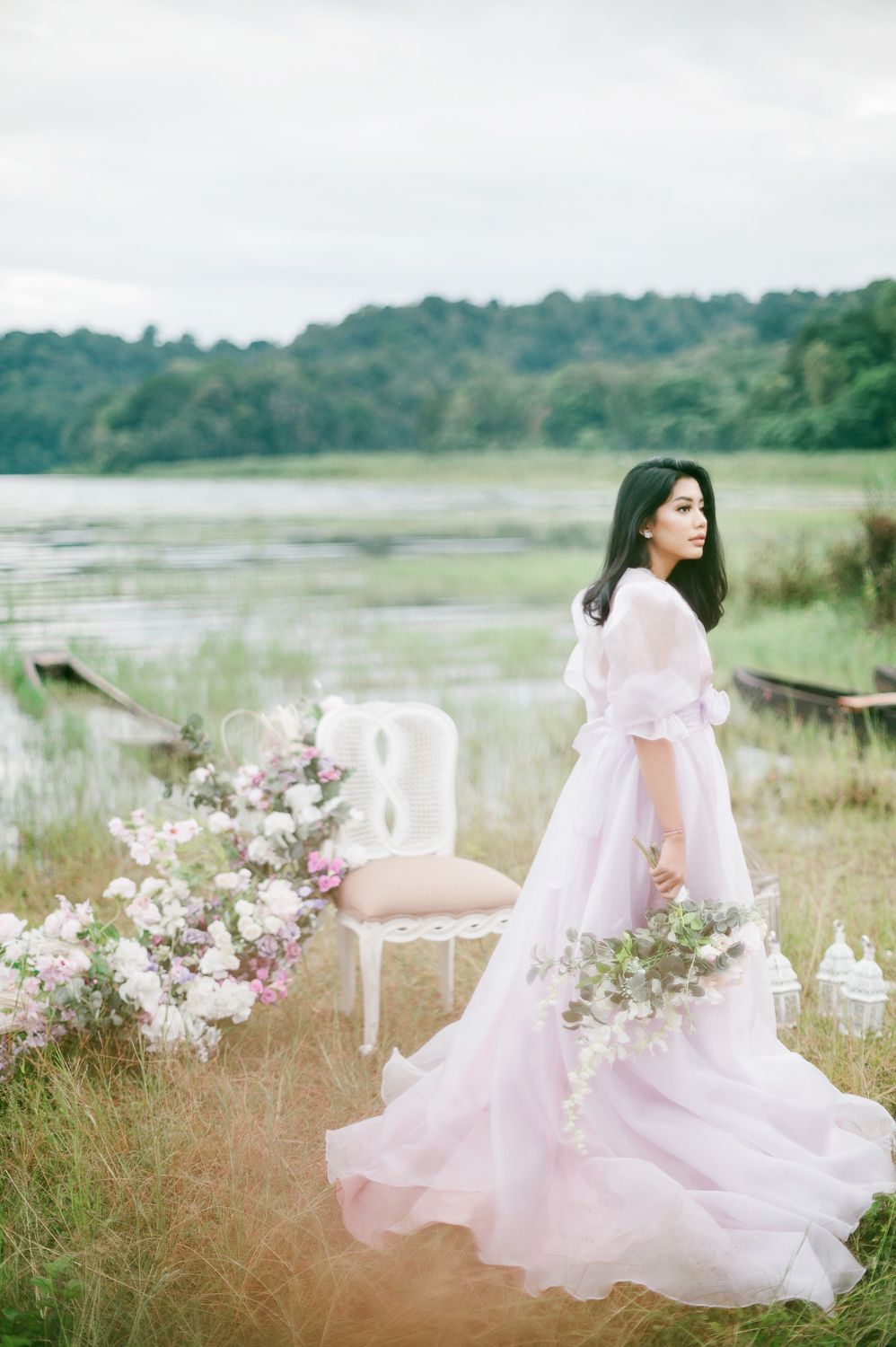 Bride in a flowing pink gown stands by a lakeside meadow surrounded by floral arrangements and a white chair.