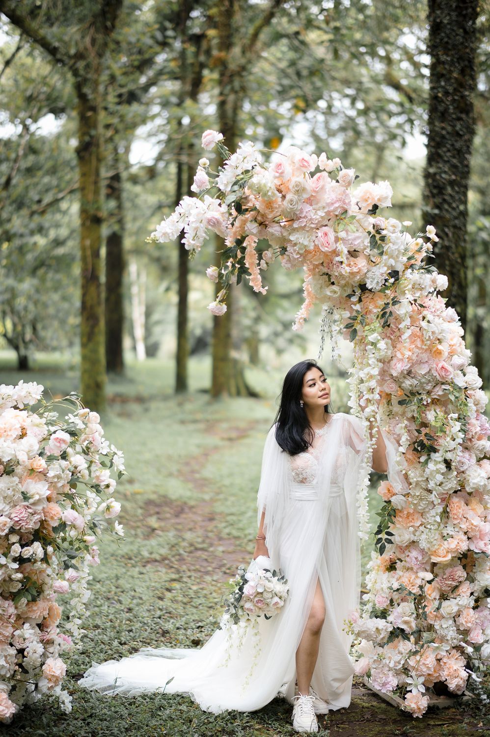 Bride in white dress standing under a floral arch with blush pink flowers in a lush green forest setting.