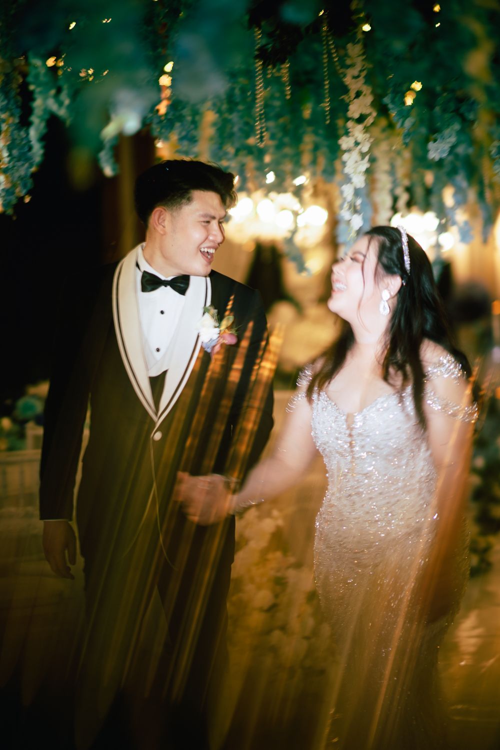 A joyful couple shares a laugh during their wedding reception, groom in tuxedo and bride in beaded gown, surrounded by lush greenery.