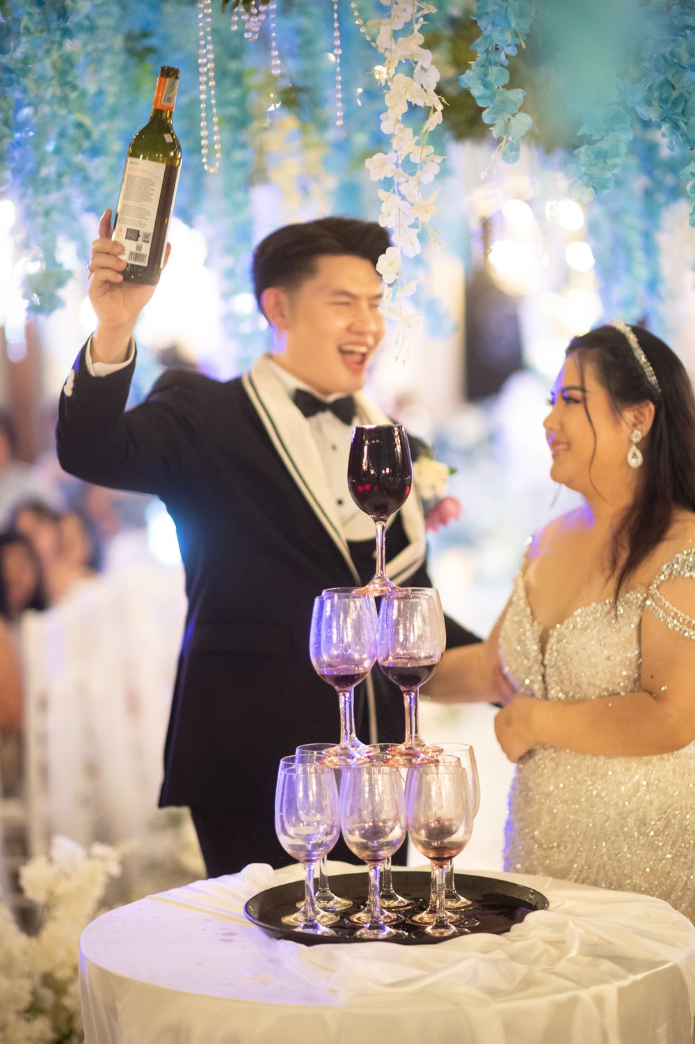 Smiling groom in tuxedo holds wine bottle above champagne tower as bride in silver gown looks on at wedding reception.