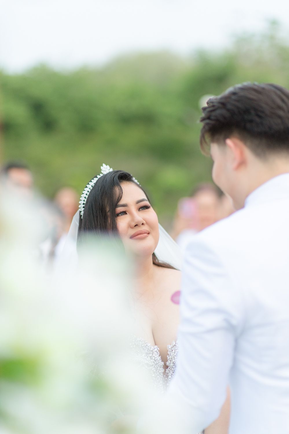 Bride in a beaded gown and tiara gazes emotionally at groom during an outdoor wedding ceremony.