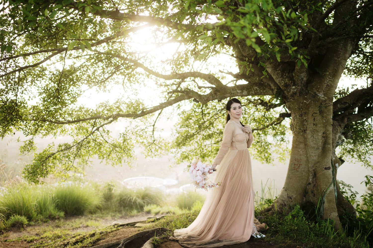 Bride in blush pink gown poses gracefully under a large tree in a lush garden setting with soft natural light.