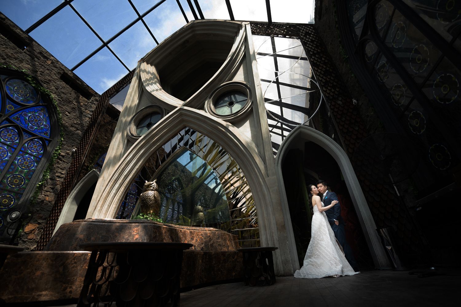 Bride and groom share a romantic kiss in a stunning architectural wedding venue with gothic arches and glass ceiling.