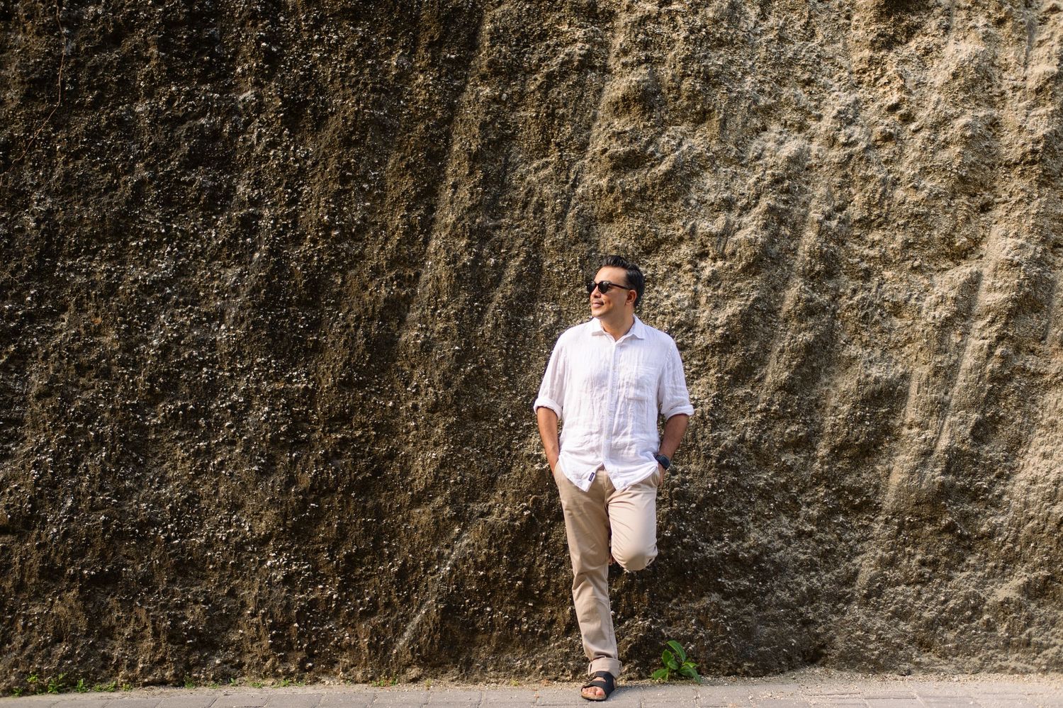 Man in white shirt and khaki pants leaning against a rugged rock wall outdoors wearing sunglasses.