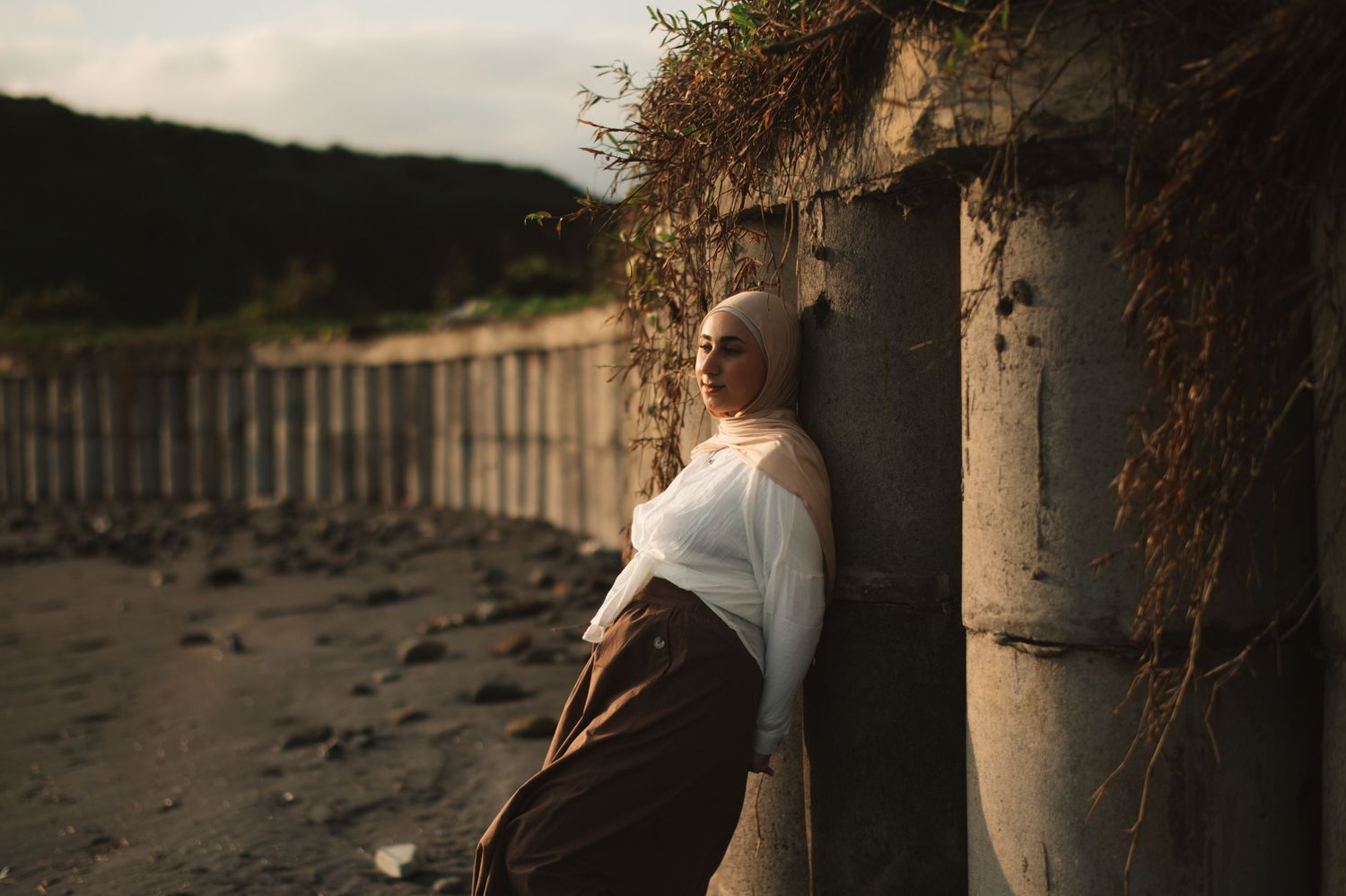 A woman in a white top and brown skirt leans against a concrete wall outdoors at golden hour.