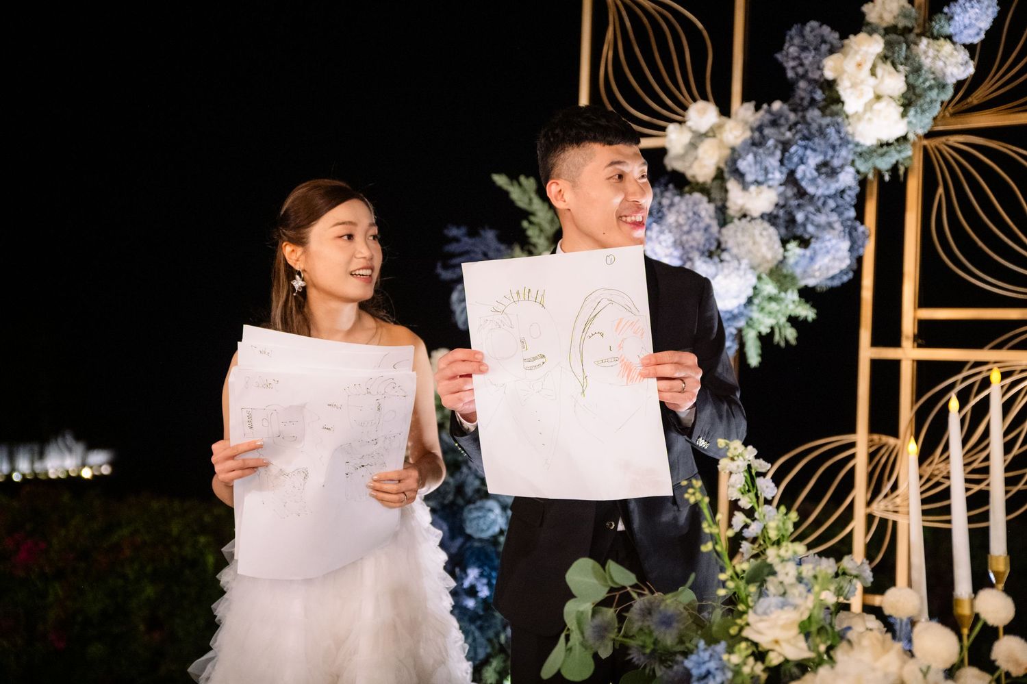 Smiling bride and groom hold up hand-drawn sketches at an evening wedding reception surrounded by blue and white floral arrangements.