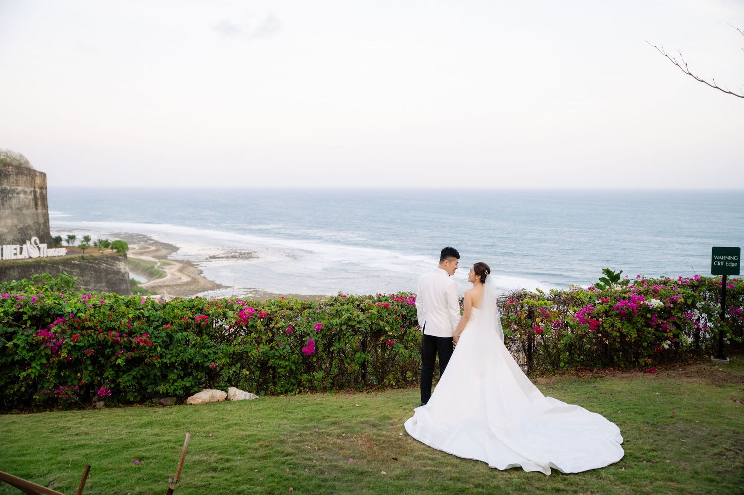 Bride in a long white gown and groom in a white shirt stand on a grassy cliffside garden with pink flowers overlooking the ocean.