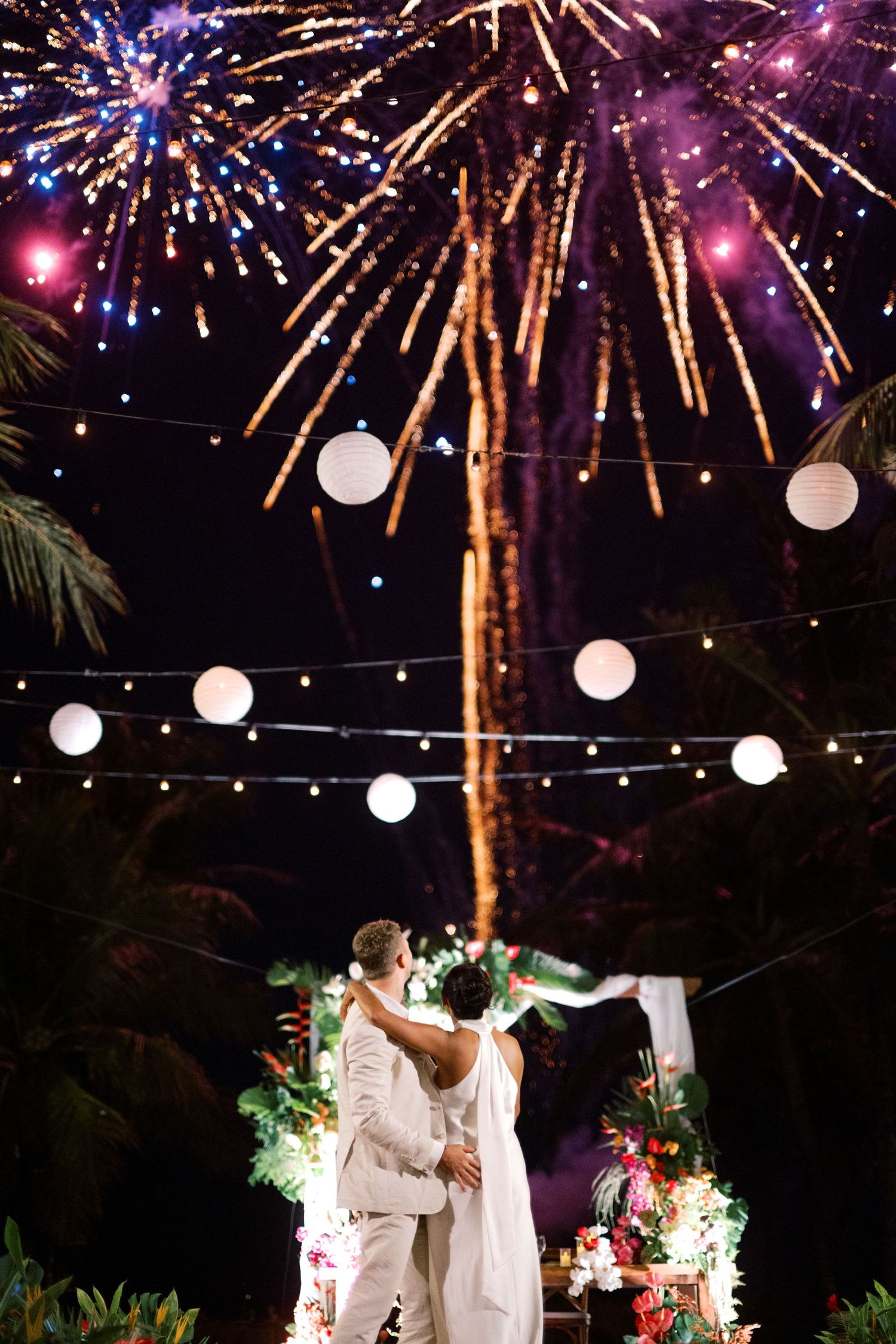 Couple embracing under colorful fireworks display at tropical outdoor wedding with string lights and paper lanterns.