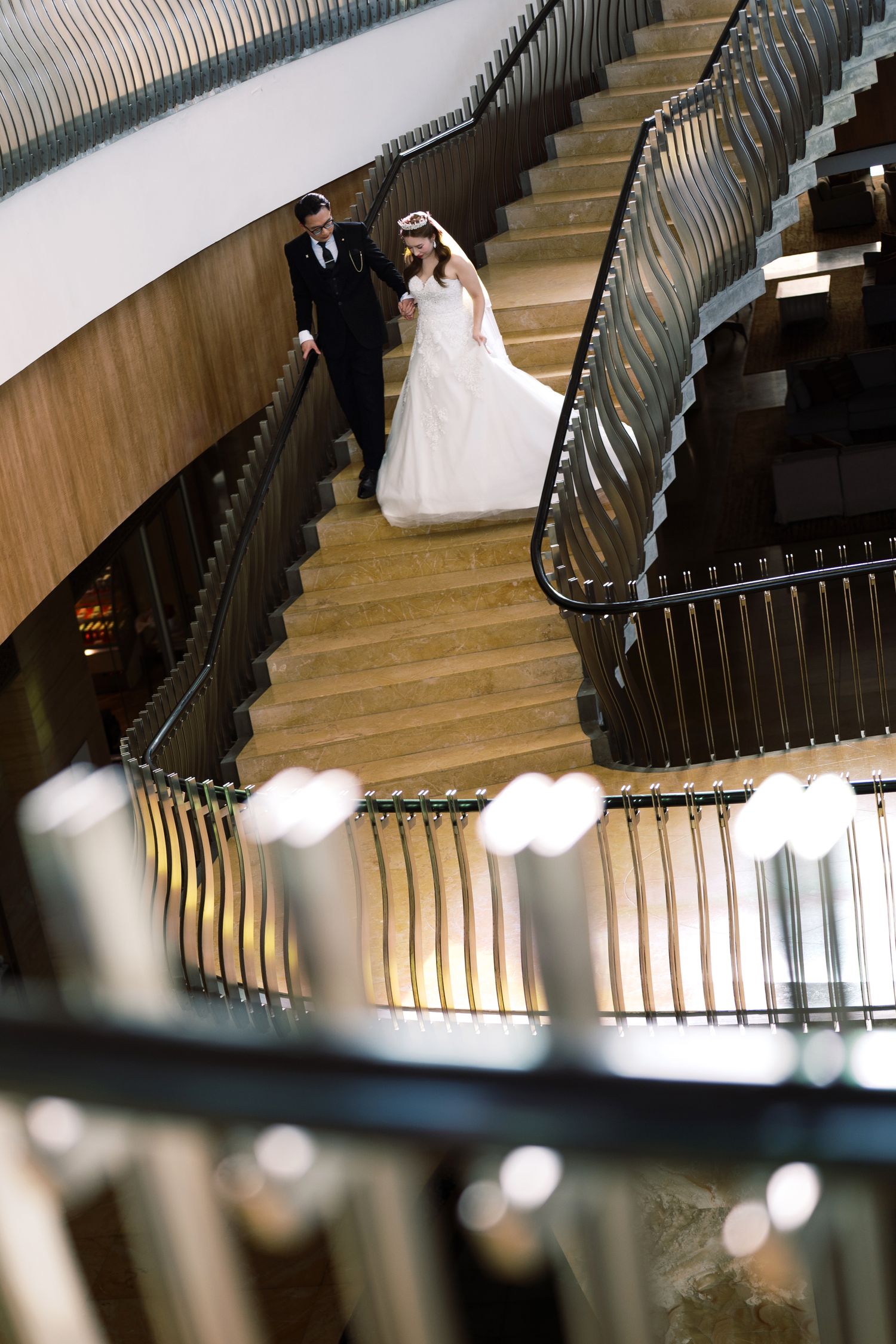 Bride and groom descend elegant curved staircase in modern venue, bride in white gown, groom in black suit.