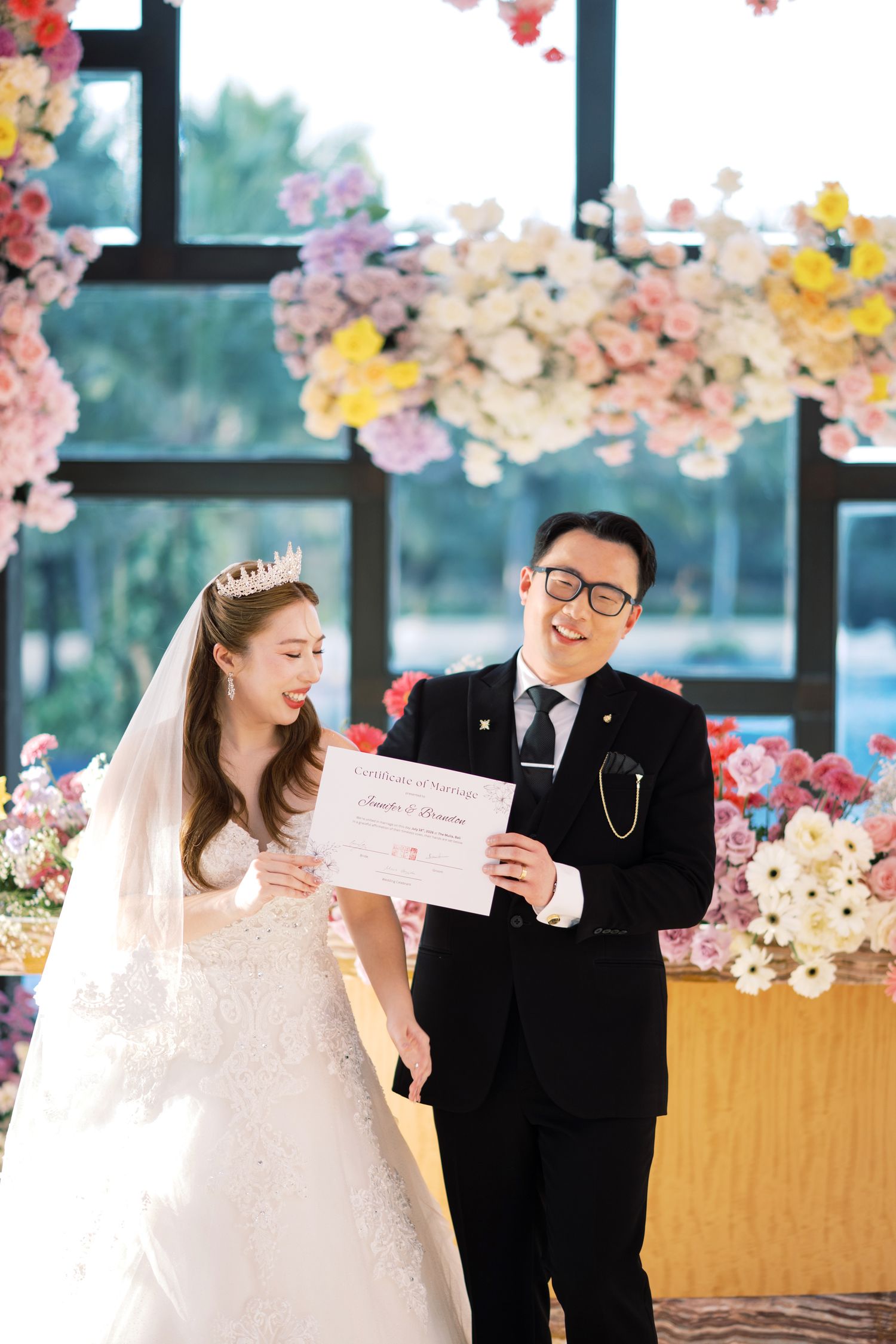 Bride in white gown and groom in black suit holding wedding certificate at floral ceremony backdrop.