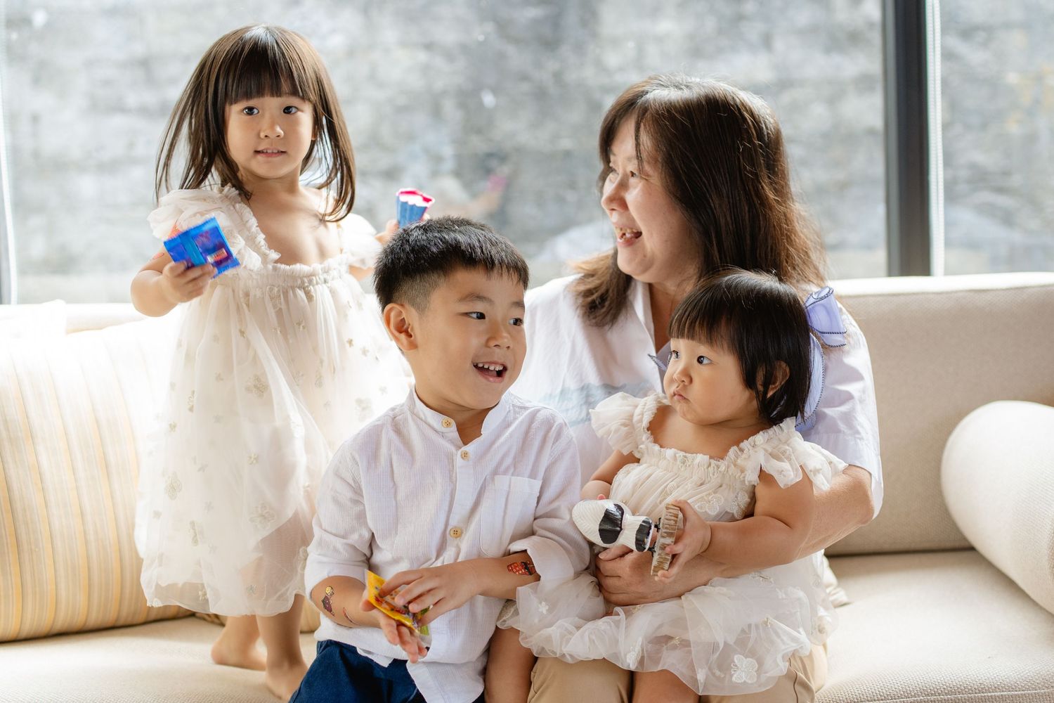A smiling Asian mother sits on a sofa with her three young children, playing with colorful toys in a bright living room.