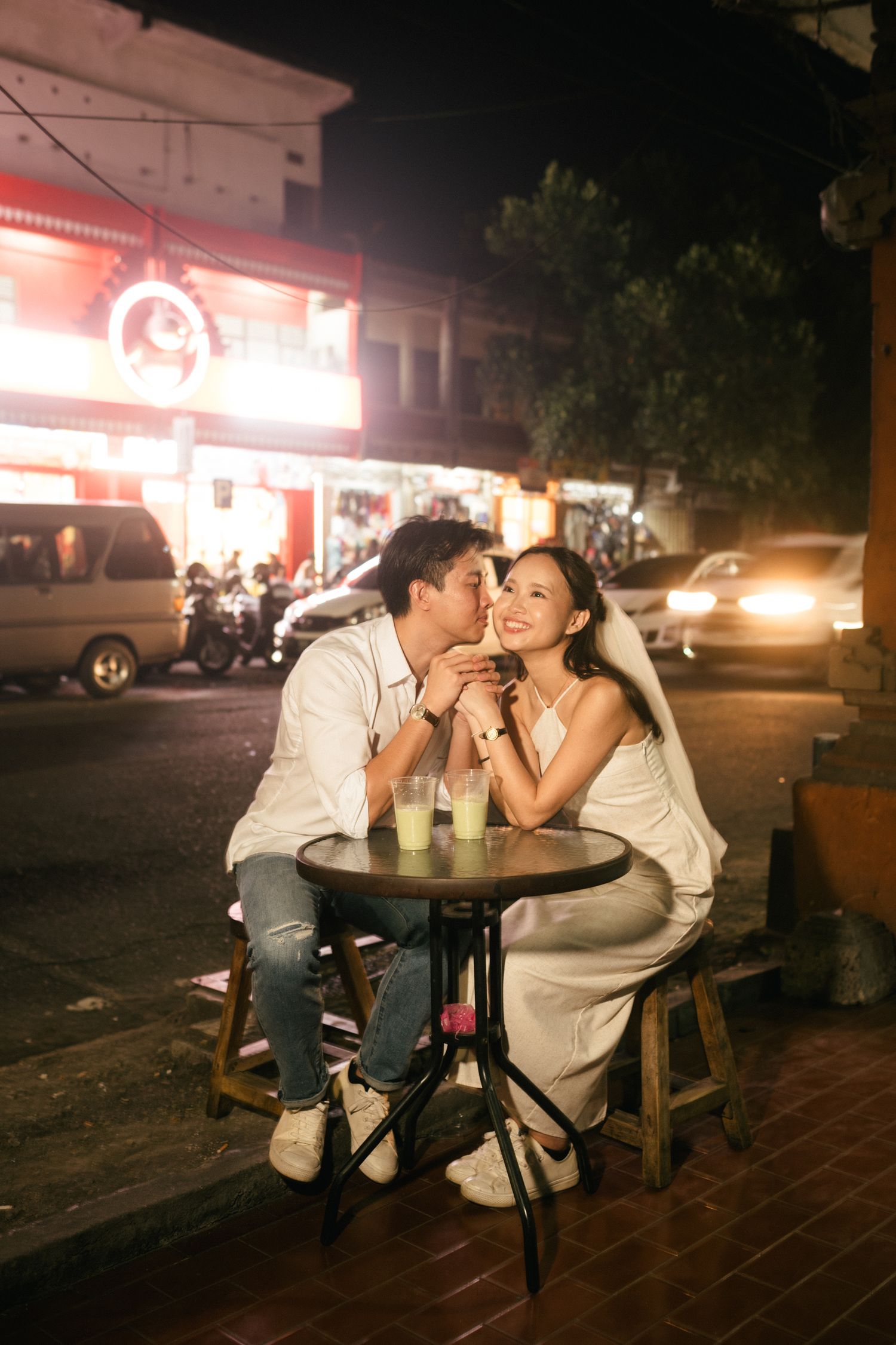 Couple in wedding attire sharing a romantic kiss at an outdoor cafe table at night with city lights behind them.