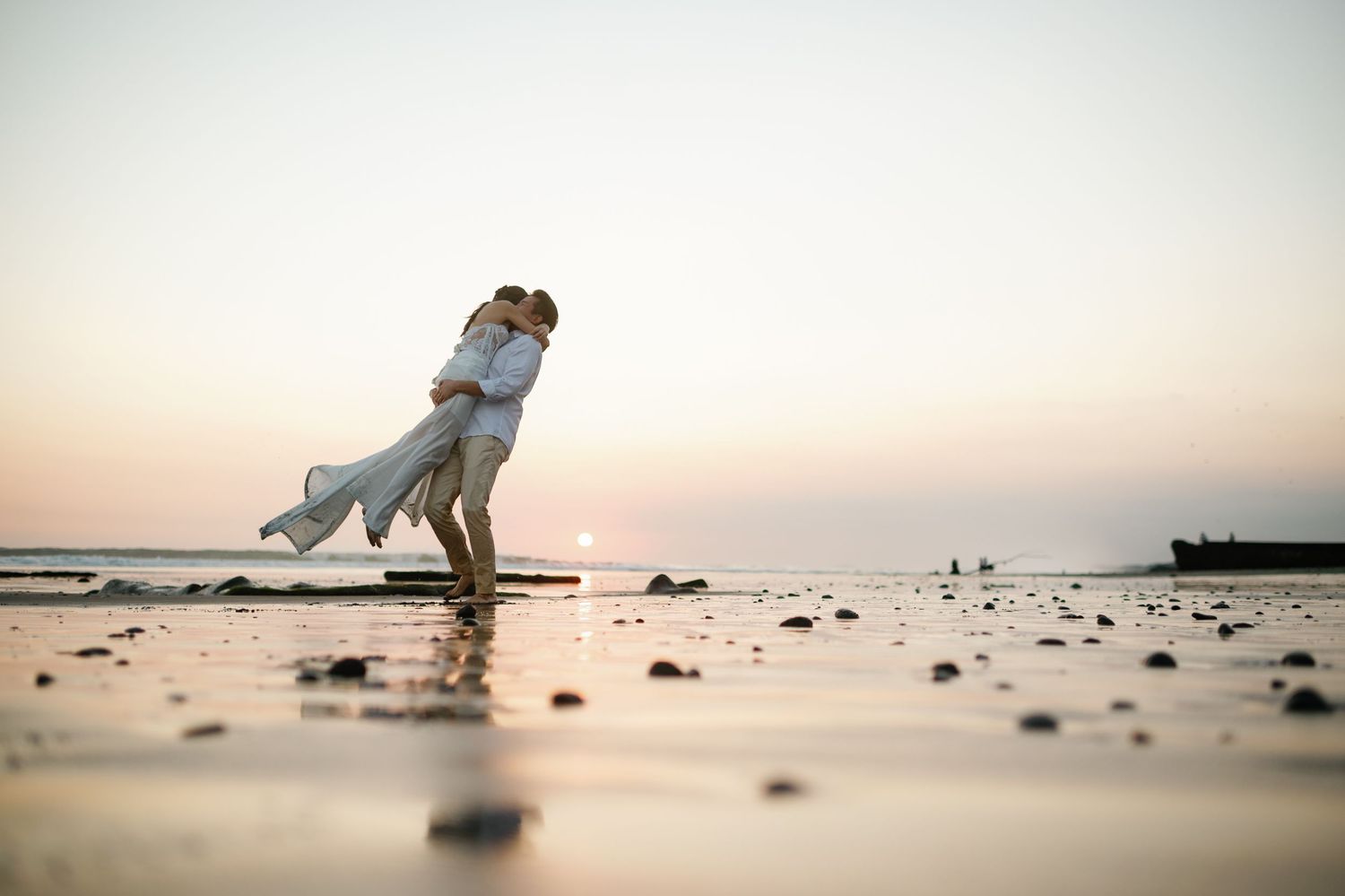 Groom lifting bride in a joyful embrace on a rocky beach during a pastel sunset.