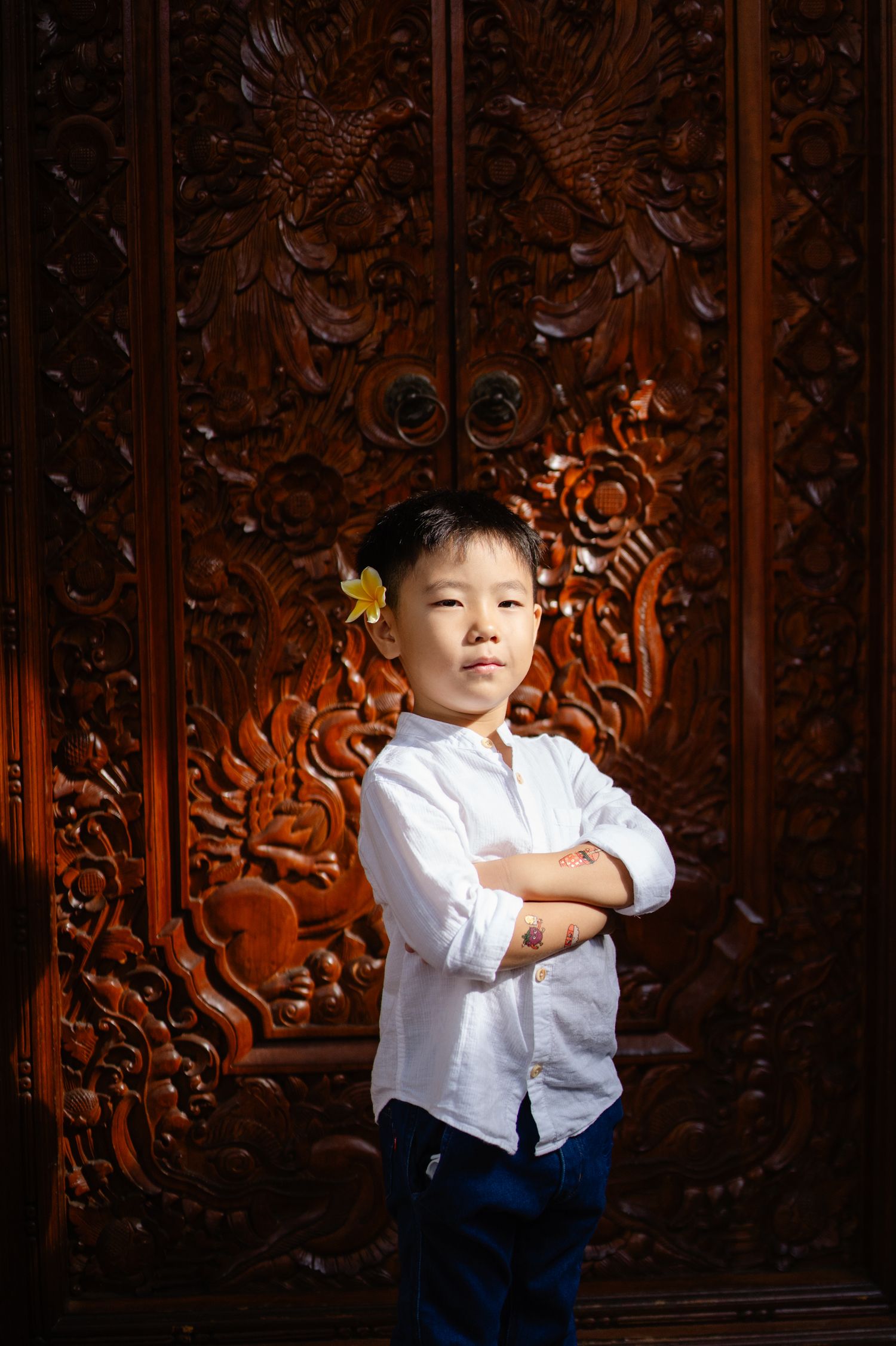 A young Asian boy in a white shirt stands confidently with arms crossed before an ornate carved wooden door.