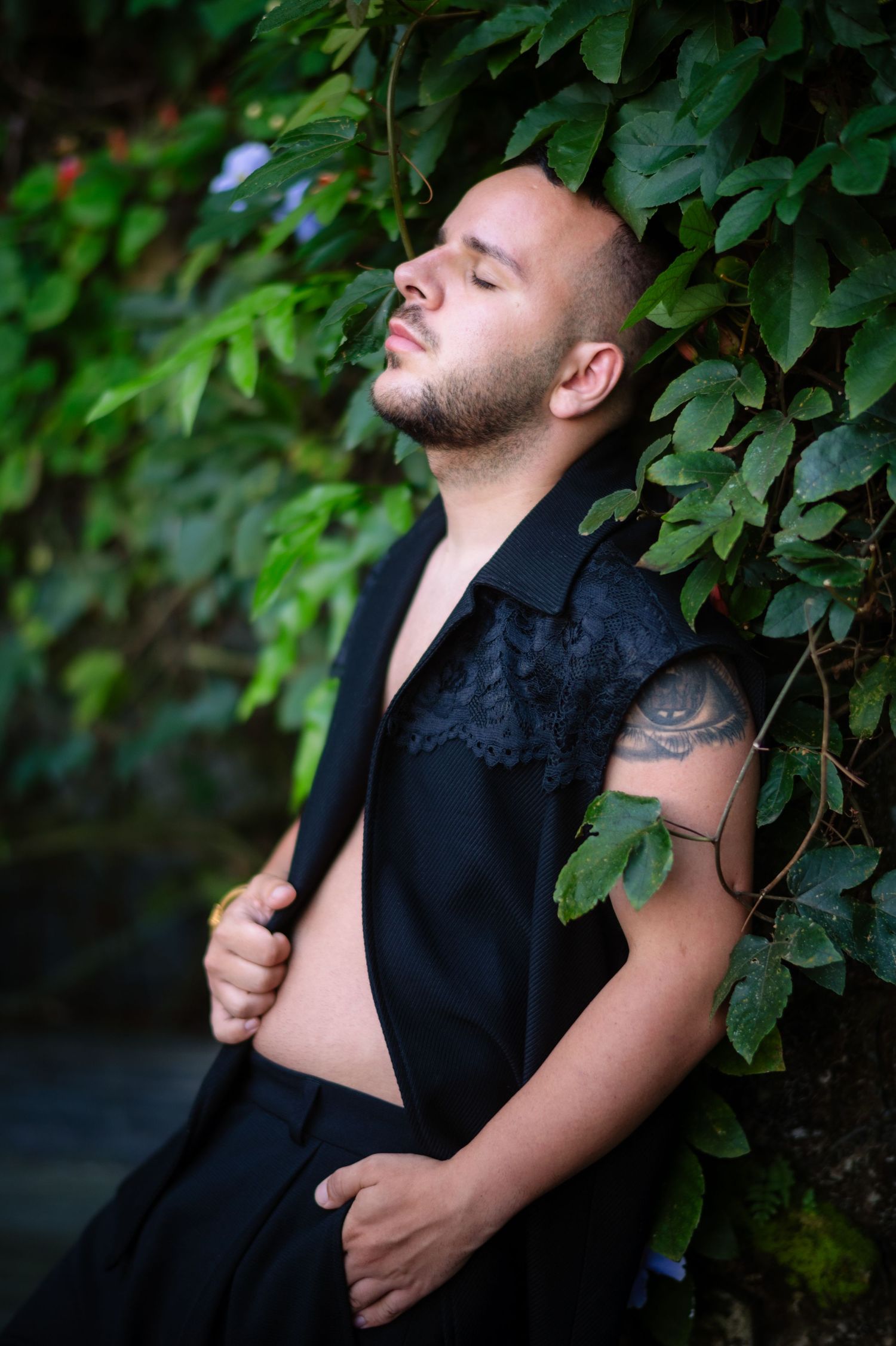 Man with tattoo leaning against ivy wall, eyes closed, wearing open black shirt outdoors.