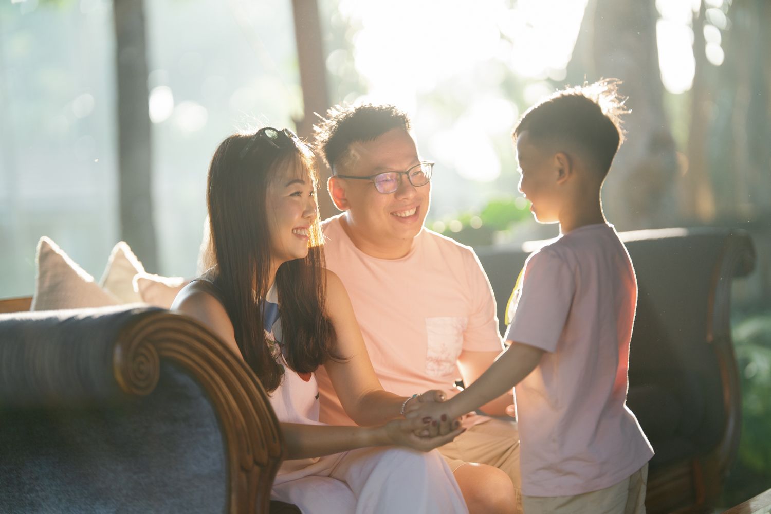 Happy Asian family with parents and young son sharing a warm moment together on a couch in sunlit room.