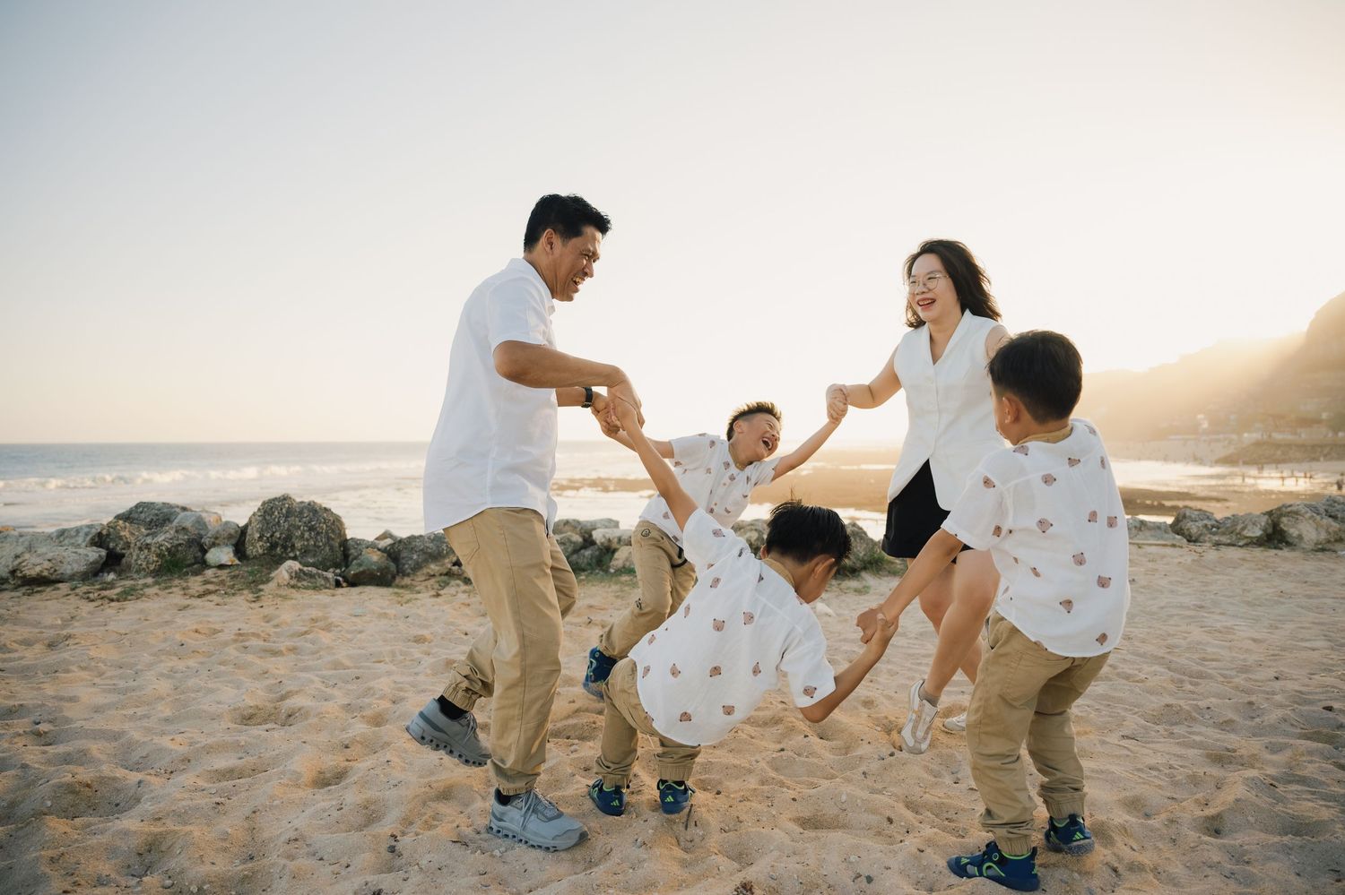 Family of five joyfully playing on a sunny beach, with parents swinging children in a circle near rocky shores.