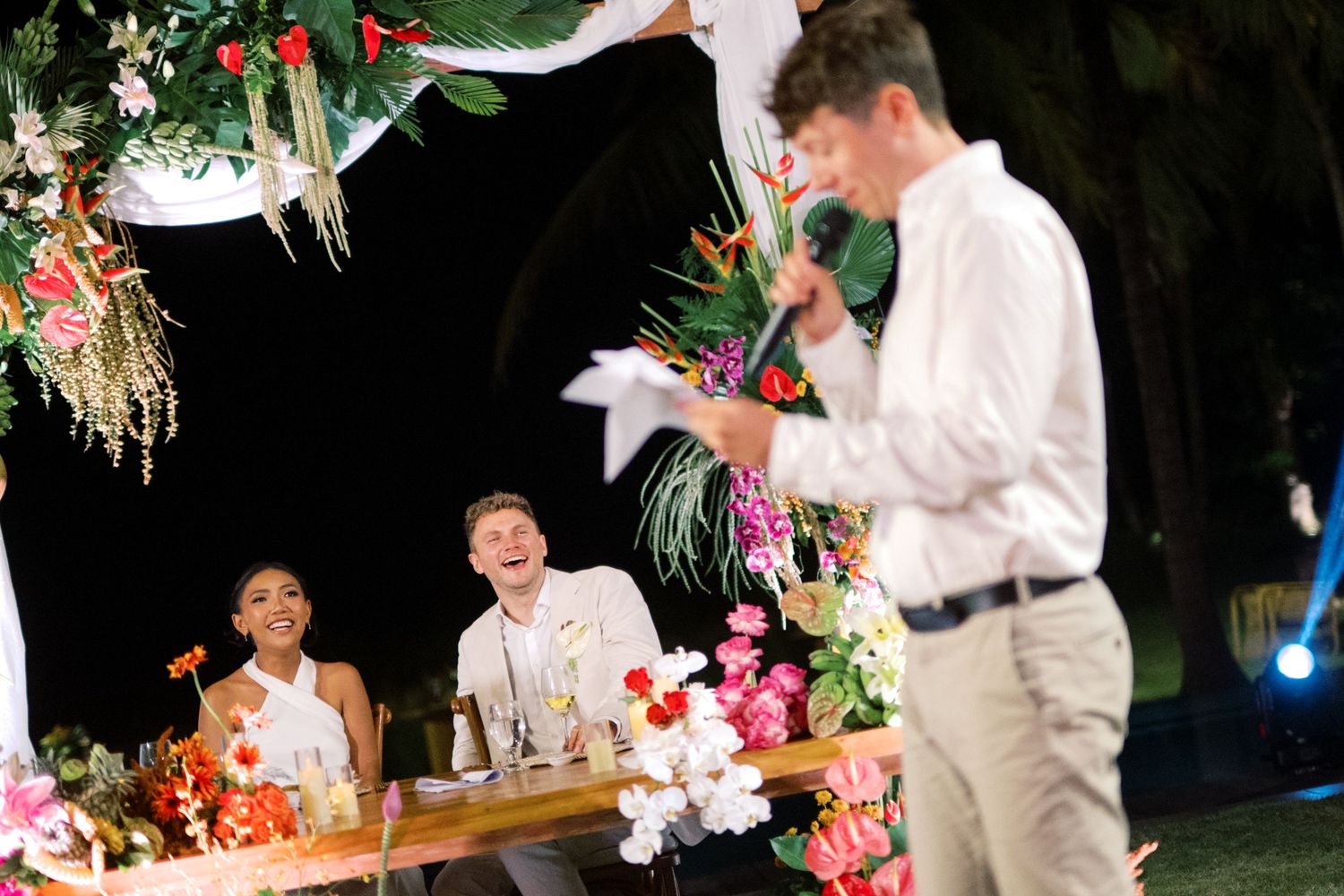 Groom laughs as groomsman delivers humorous wedding speech at floral-decorated outdoor tropical reception table.