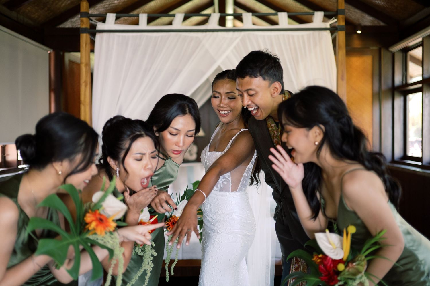 Bride and bridesmaids in green dresses share joyful moment examining wedding ring indoors.