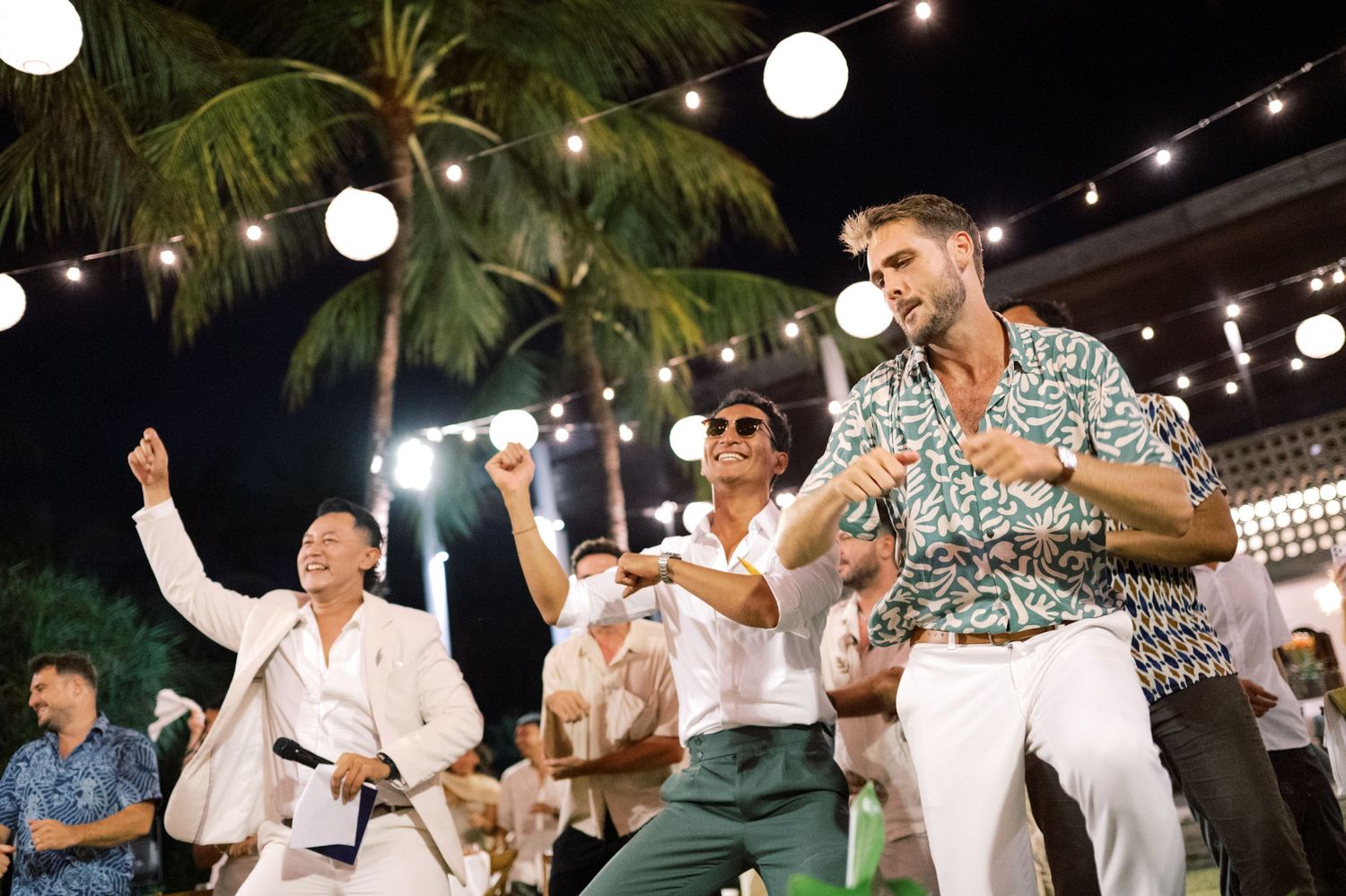 Men dance joyfully under string lights and palm trees at outdoor tropical wedding reception.