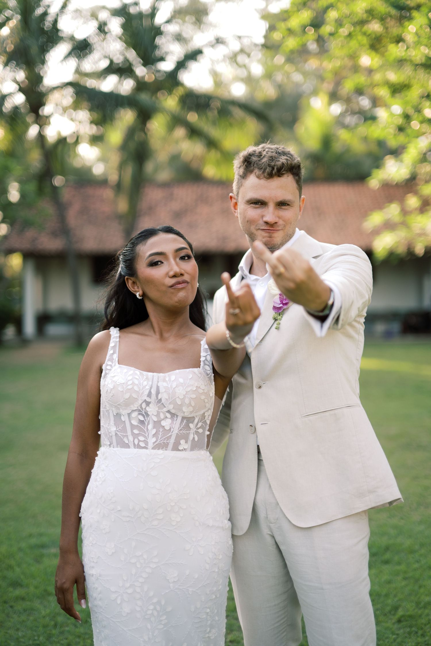 Bride and groom pointing at camera outdoors, bride in lace gown, groom in ivory suit with tropical backdrop.