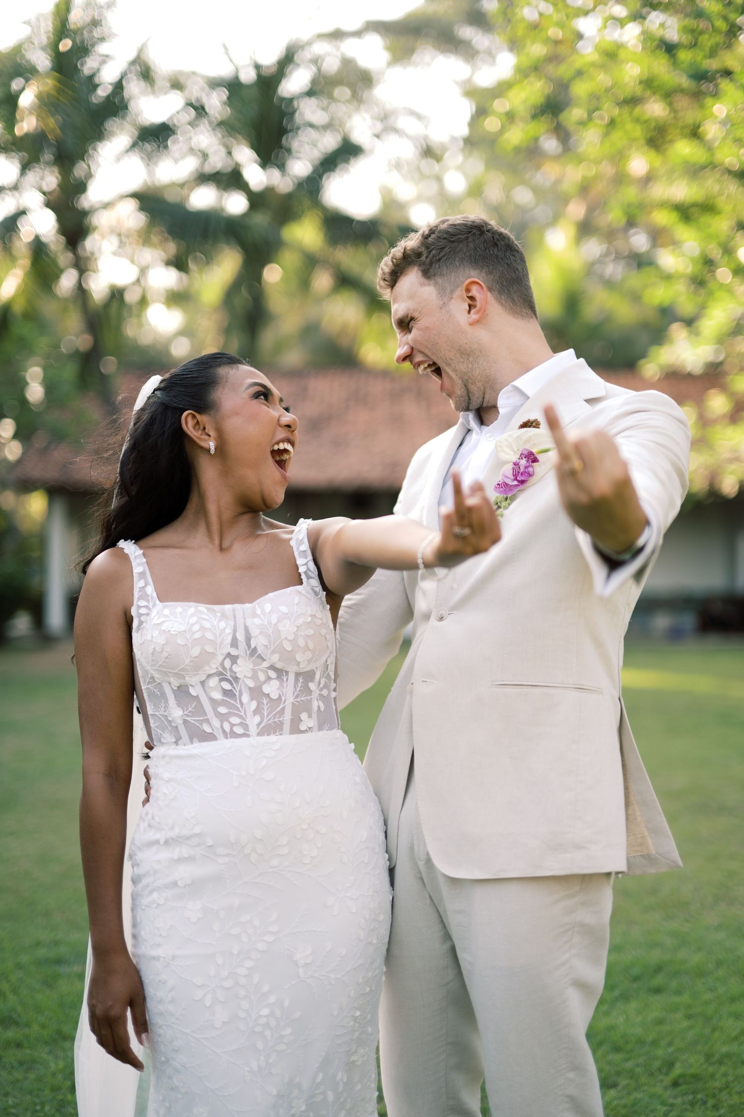 Joyful interracial couple in wedding attire laughing outdoors, bride in white lace gown, groom in beige suit with floral boutonniere.
