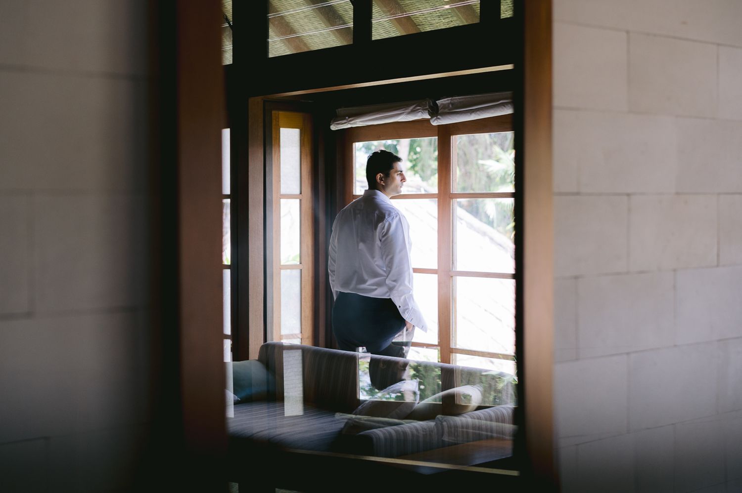 Man in white shirt standing by large windows in a serene interior space, gazing outside thoughtfully.