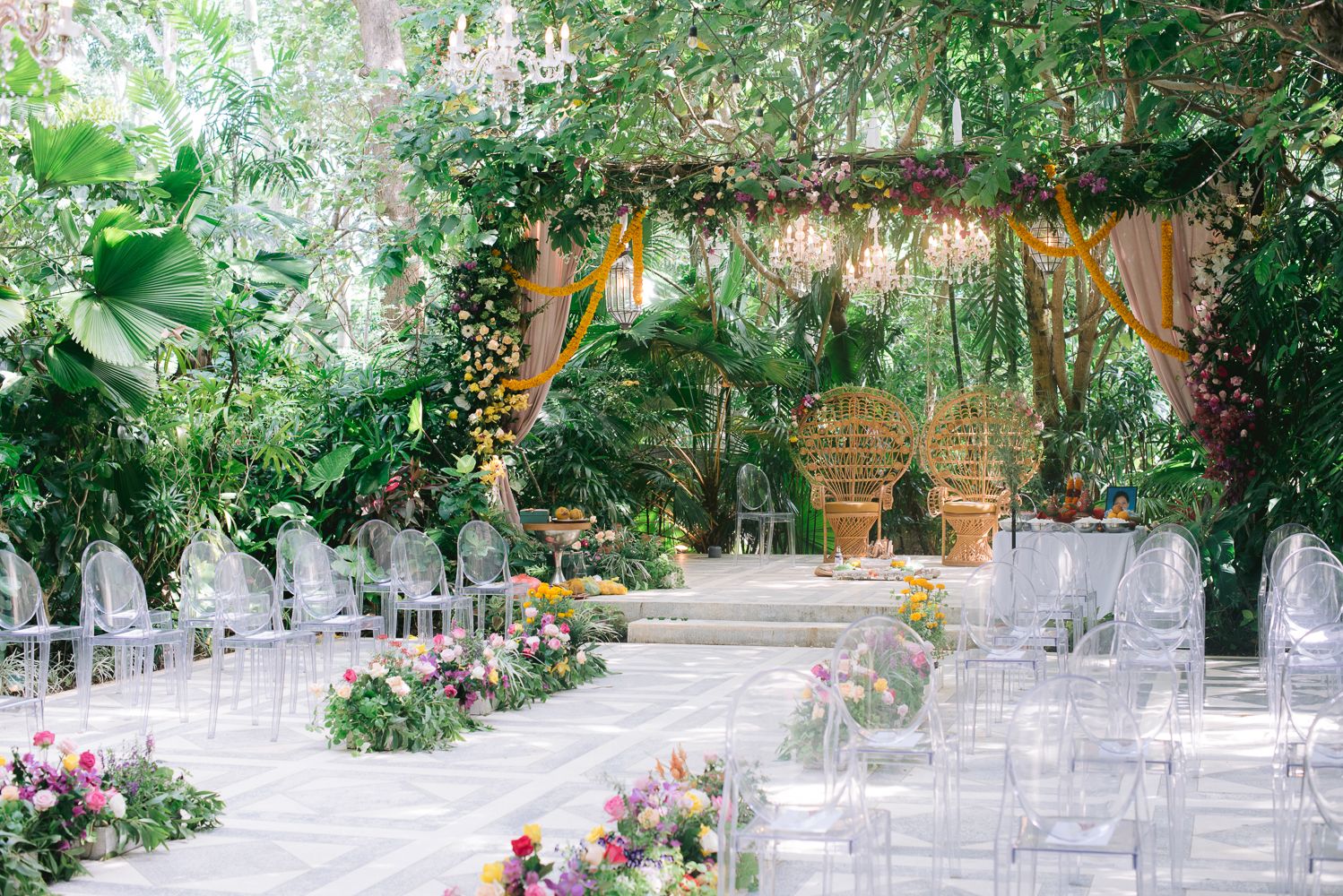 Lush garden wedding ceremony with ghost chairs, tropical greenery, colorful floral aisle, and elegant chandelier backdrop.