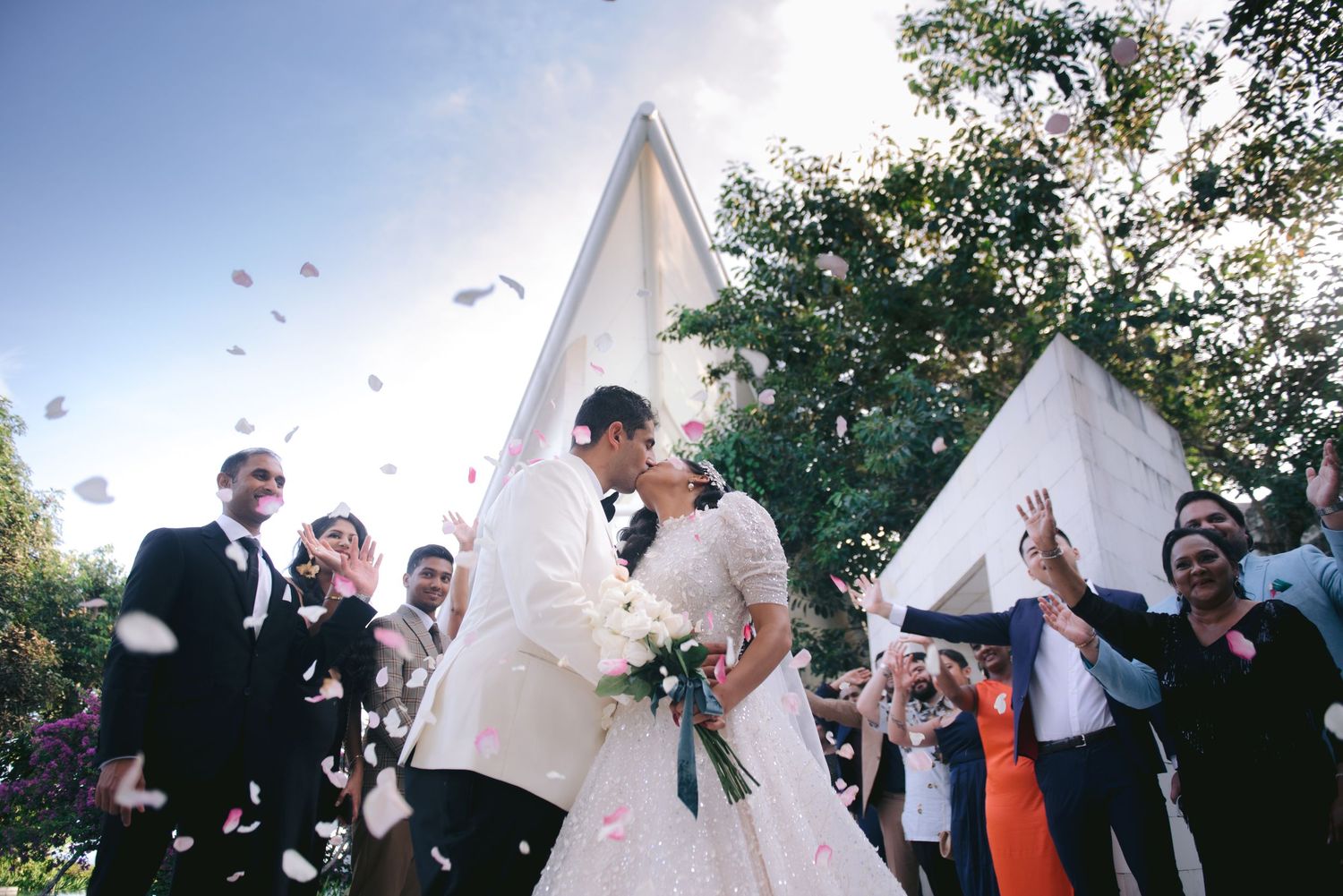 Newlyweds share a kiss as guests toss confetti outside a white chapel during a joyful outdoor wedding ceremony.