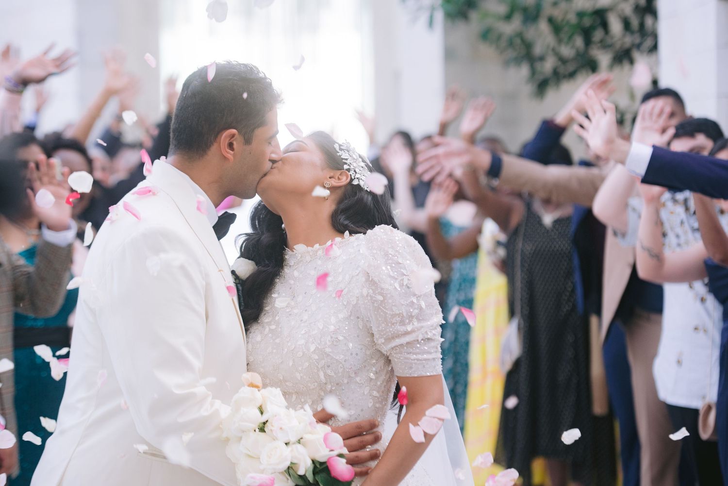 Newlywed couple sharing a kiss at wedding ceremony while guests toss rose petals in celebration.