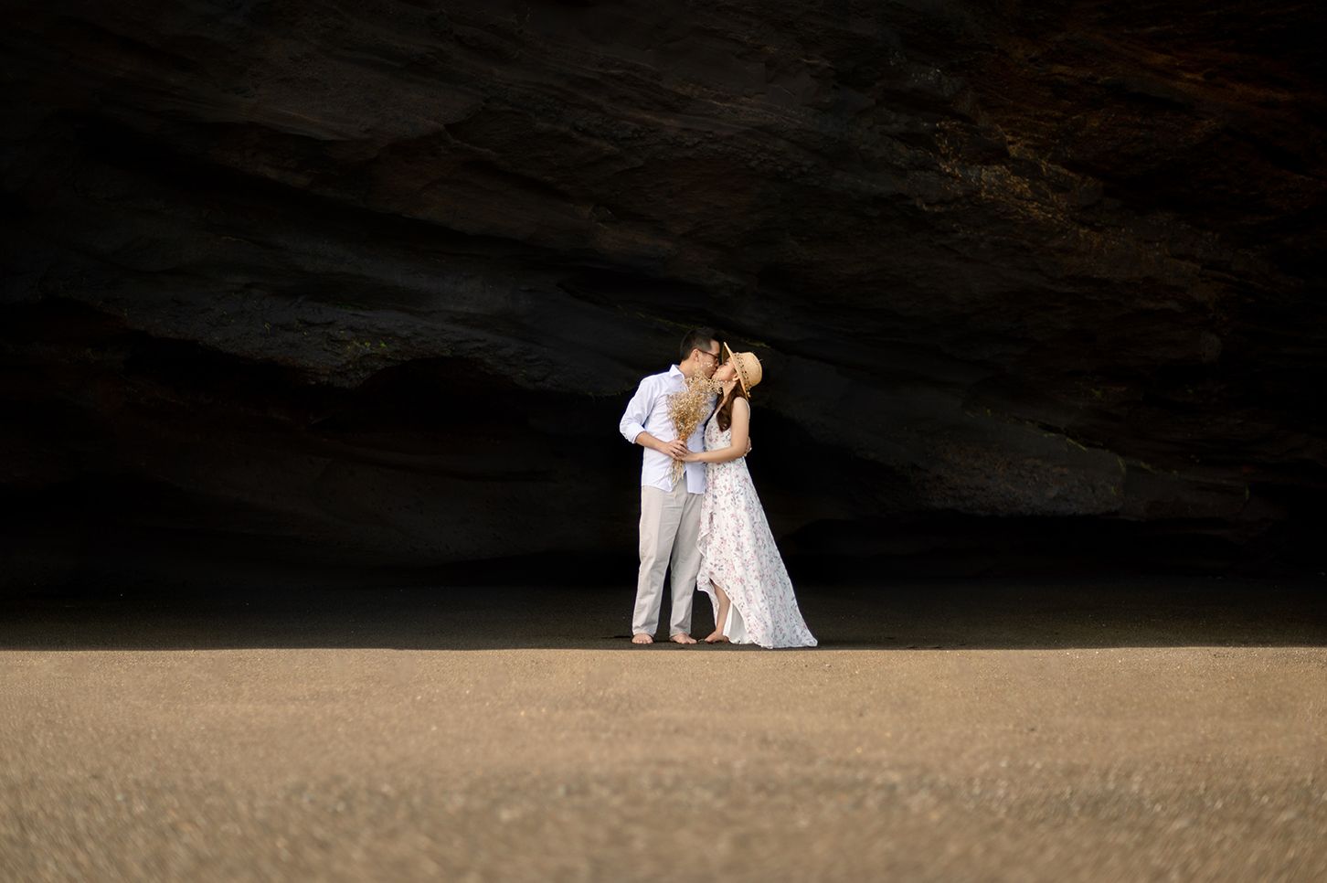 A couple shares a romantic kiss in a dramatic cave setting, bride in white gown, groom in white suit.