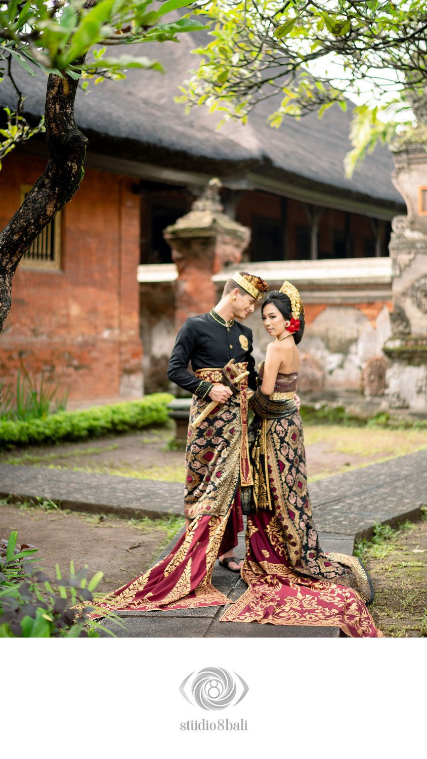 Couple in traditional Balinese wedding attire with ornate gold and red fabric, posing in a temple courtyard.