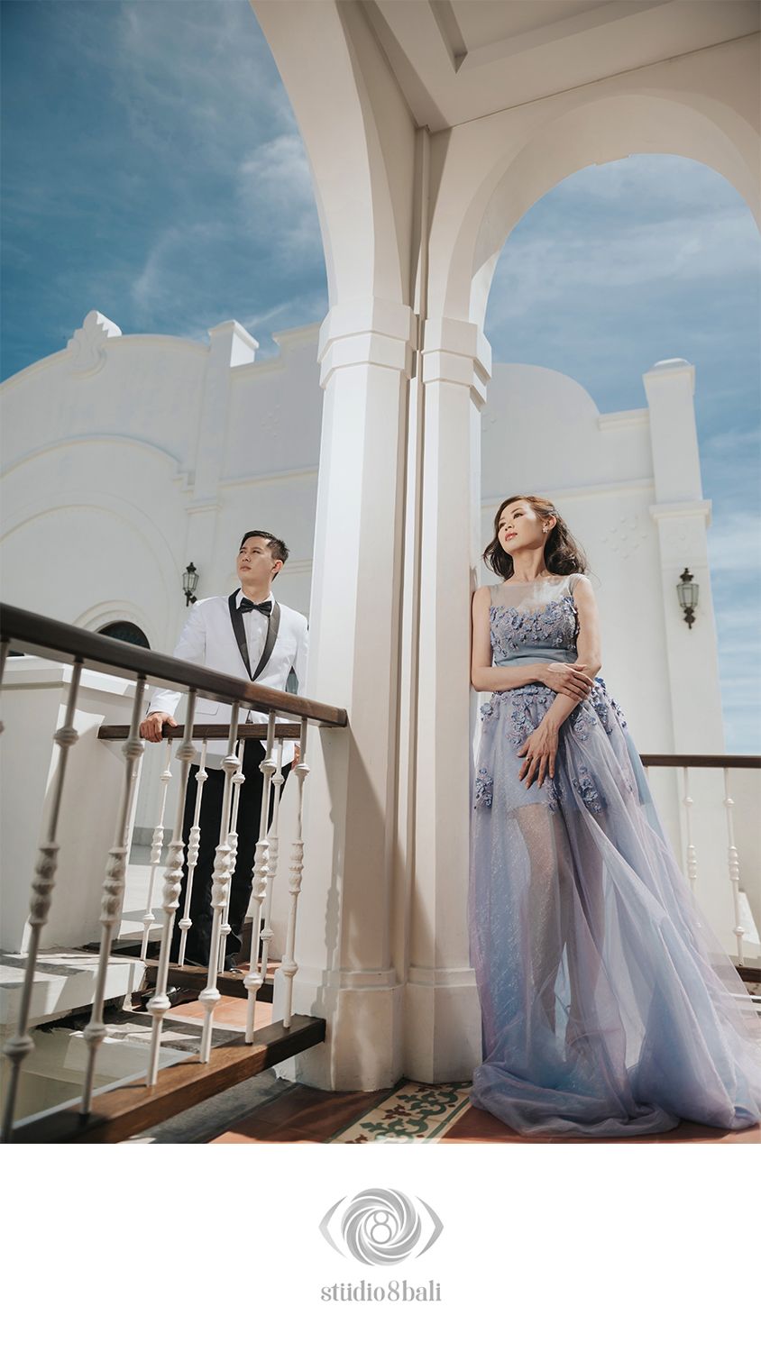 Couple in elegant attire on a white colonial balcony, woman in blue floral gown, man in white tuxedo.
