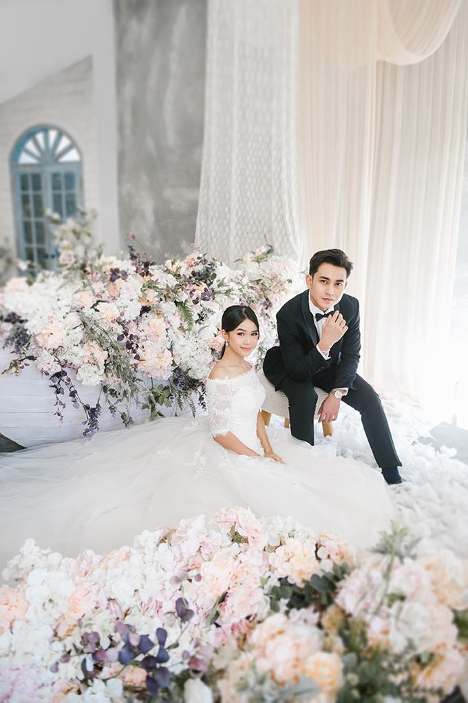 Elegant couple in wedding attire surrounded by lush pink and white floral arrangements in a bright, romantic studio setting.
