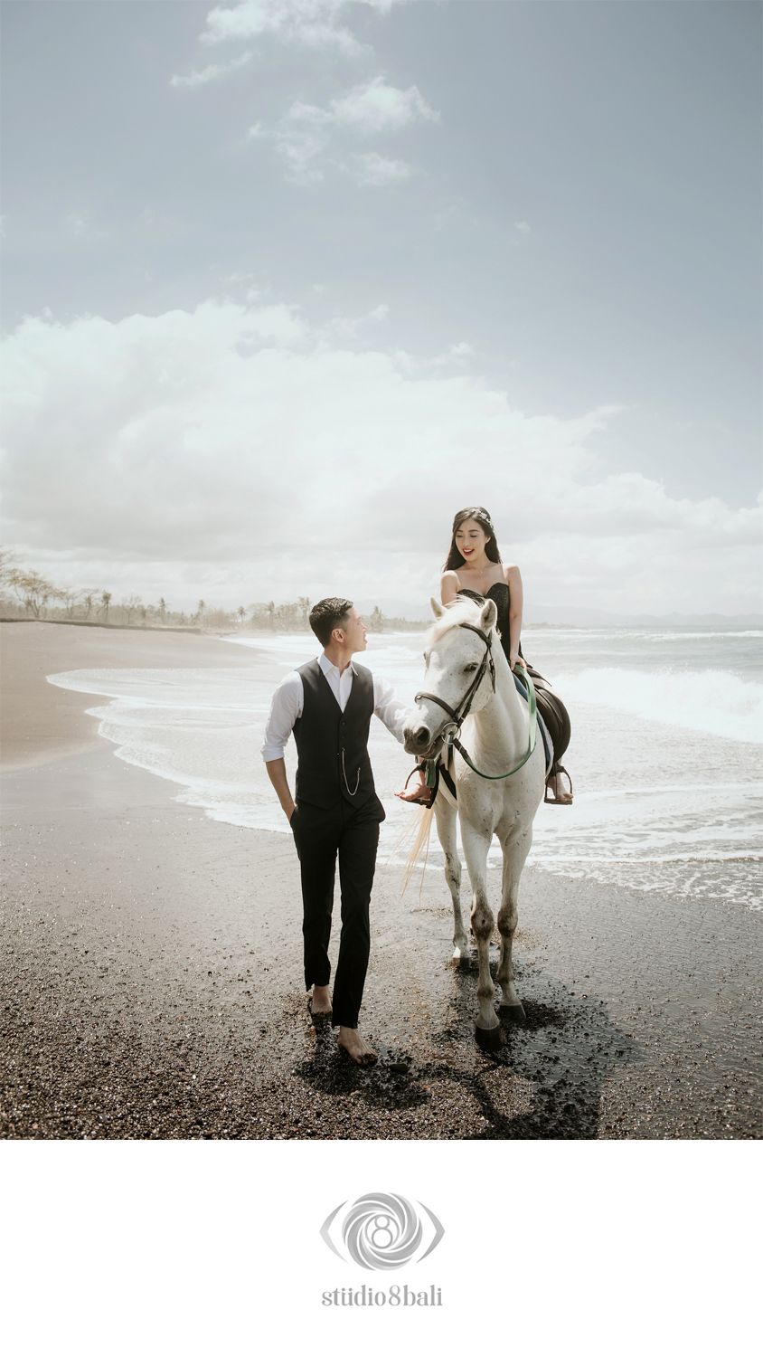 Couple in elegant attire poses on a black sand beach in Bali, woman riding a white horse, man walking alongside.